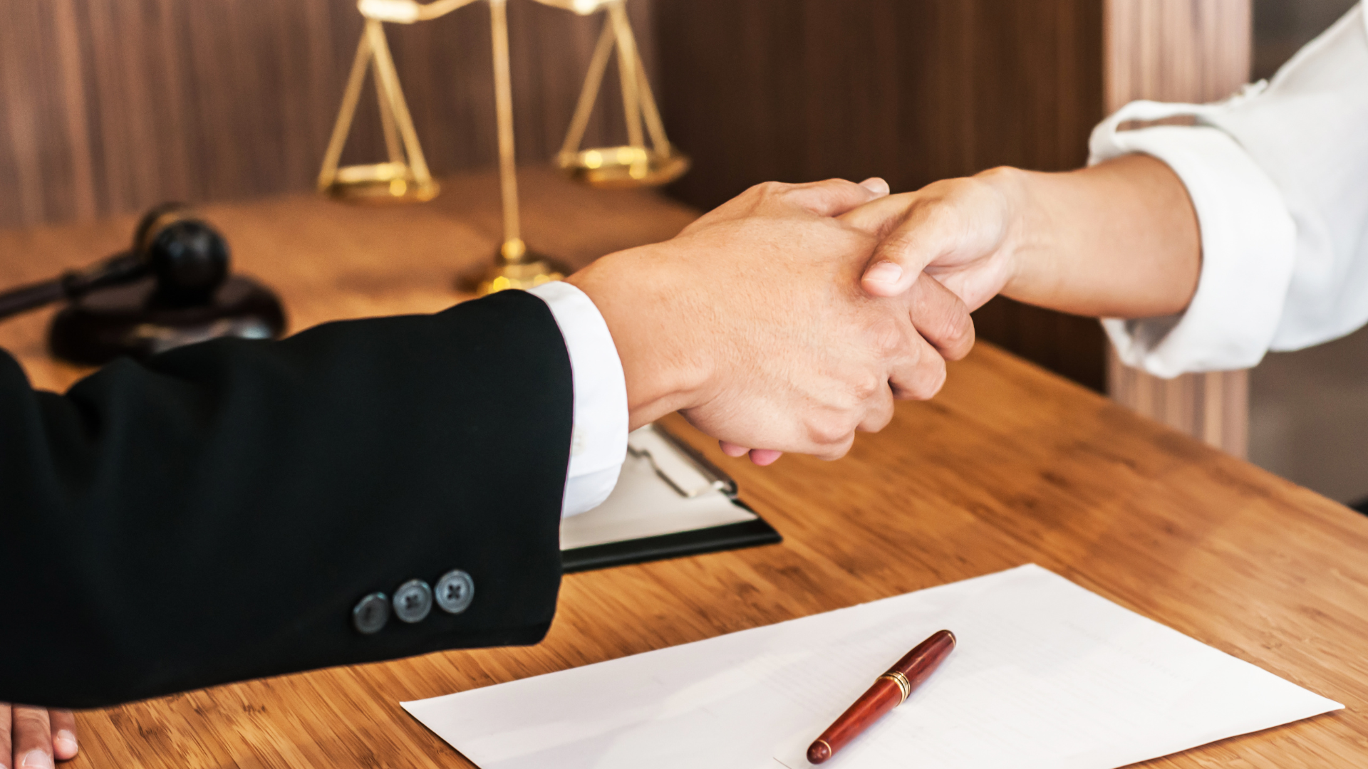 Two people shaking hands over a desk with a gavel, scales, and paperwork.