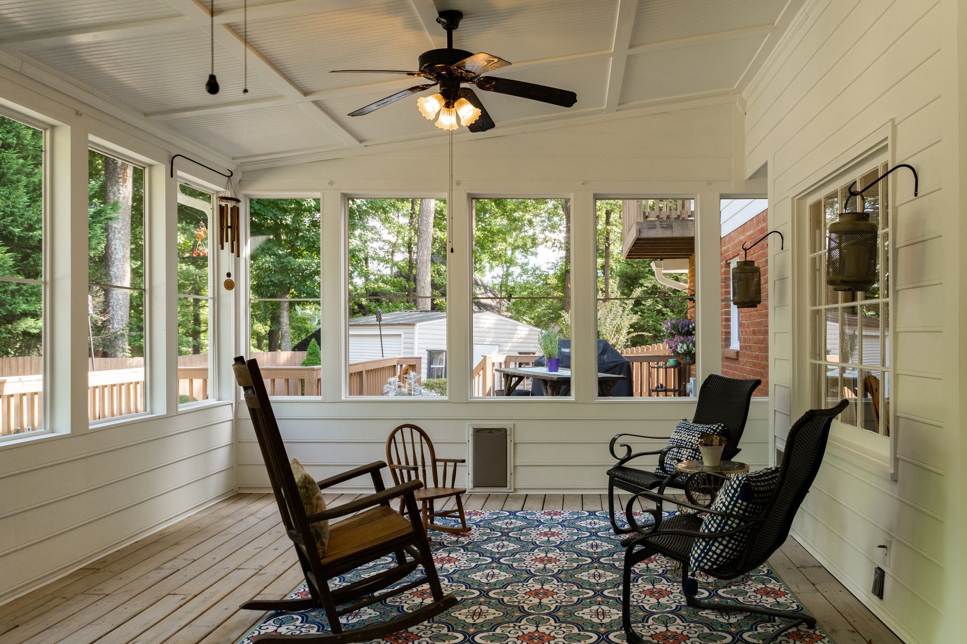 A sunroom with white walls, a ceiling fan, a patterned rug, and several chairs, including a wooden rocking chair.