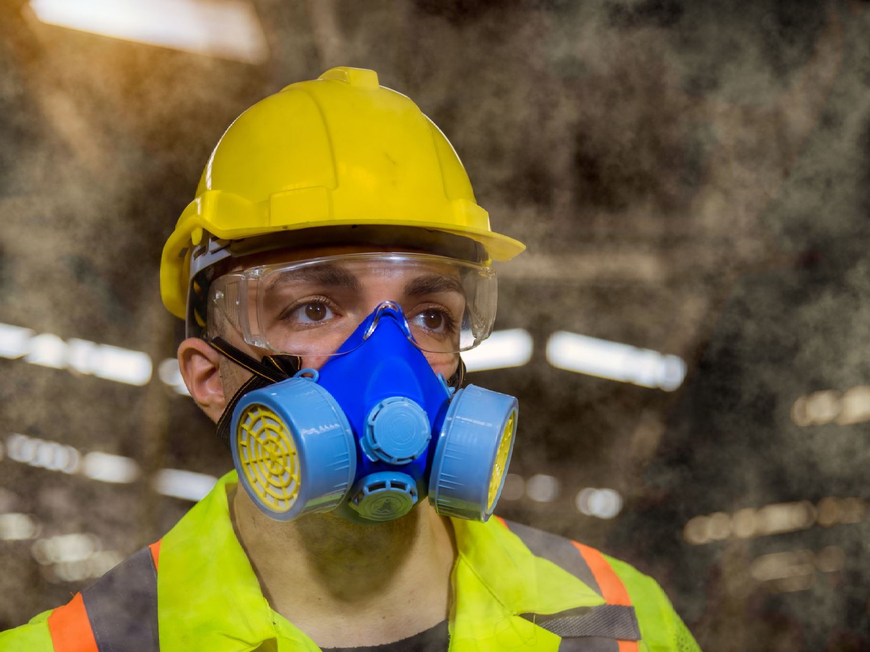 Construction worker wearing safety gear, respirator, goggles, and yellow hard hat in dusty environment.