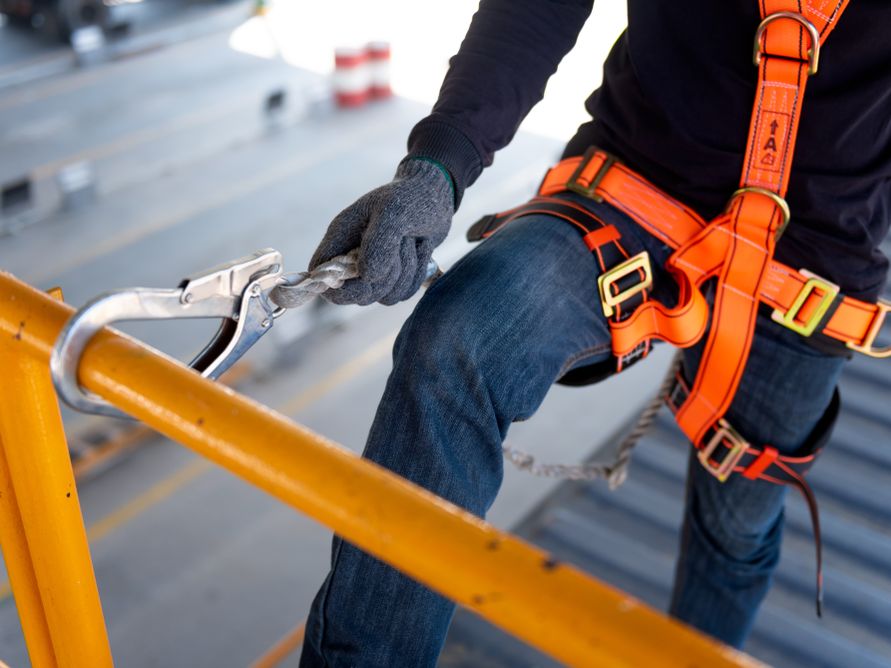 Worker in orange safety harness clipping a lanyard to a yellow handrail.