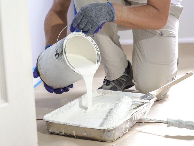 Person pours white paint from a can into a paint tray, preparing to paint.