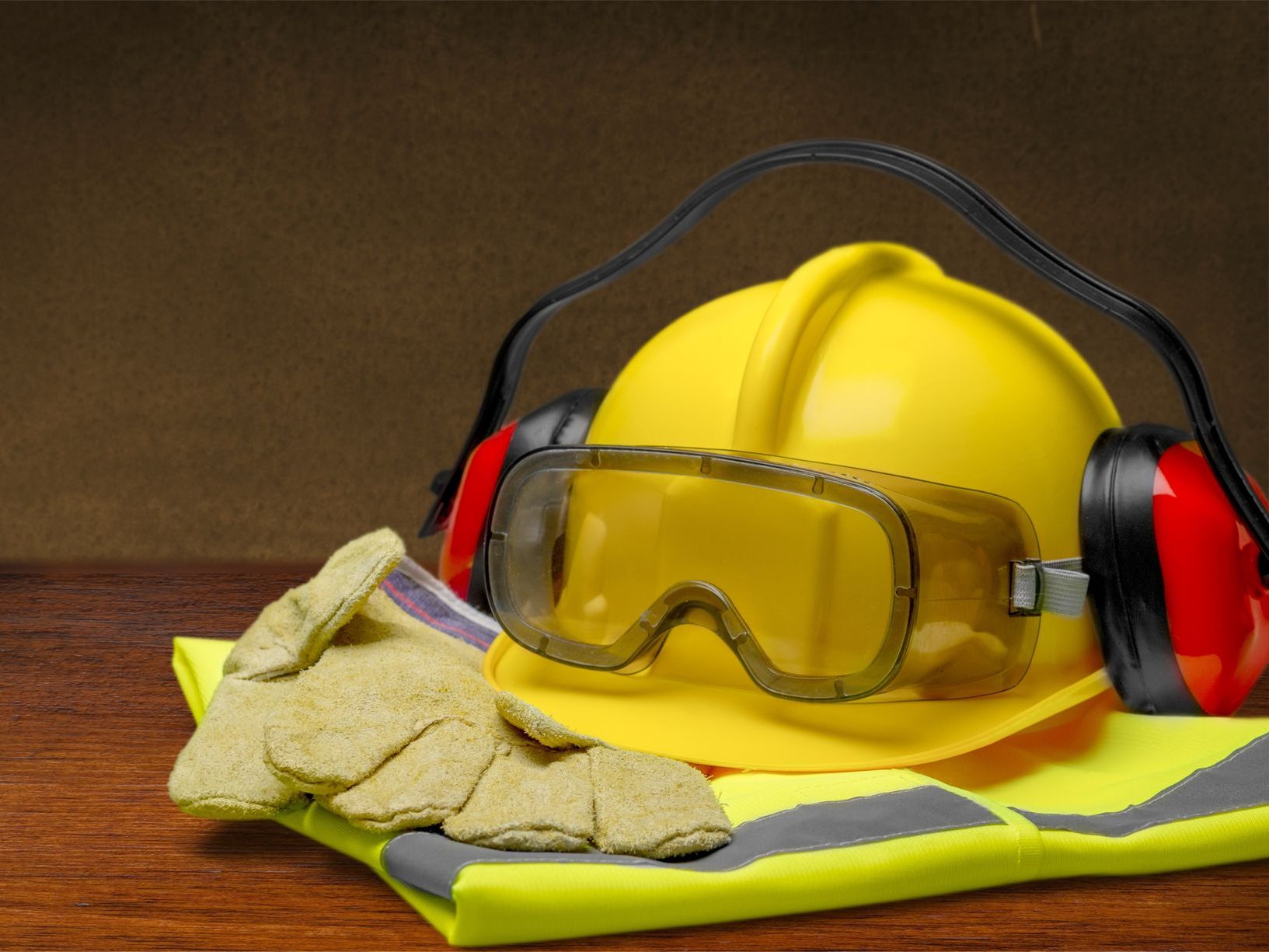 Yellow hard hat, safety glasses, ear protection, gloves, and reflective vest on a wooden surface.