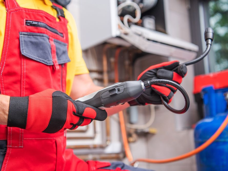 Technician in red overalls inspecting a heating system with a gas leak detector.