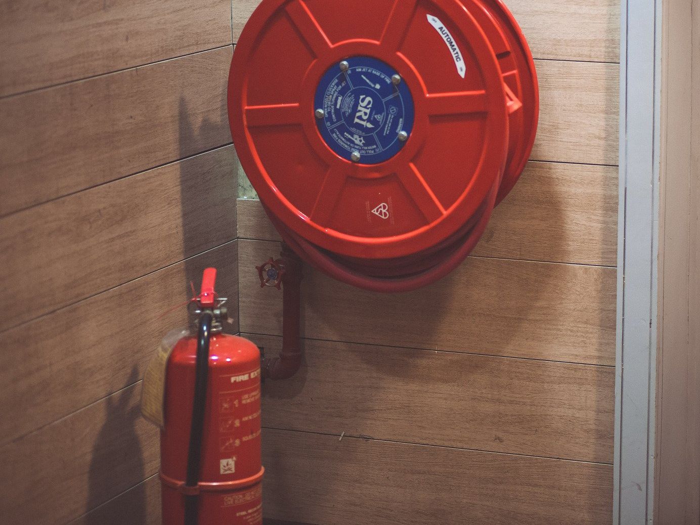 Red fire hose reel and fire extinguisher mounted in a corner against a wood-paneled wall.