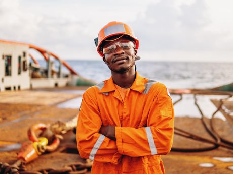 Man in orange work suit, safety glasses, and helmet on ship deck, arms crossed, smiling.