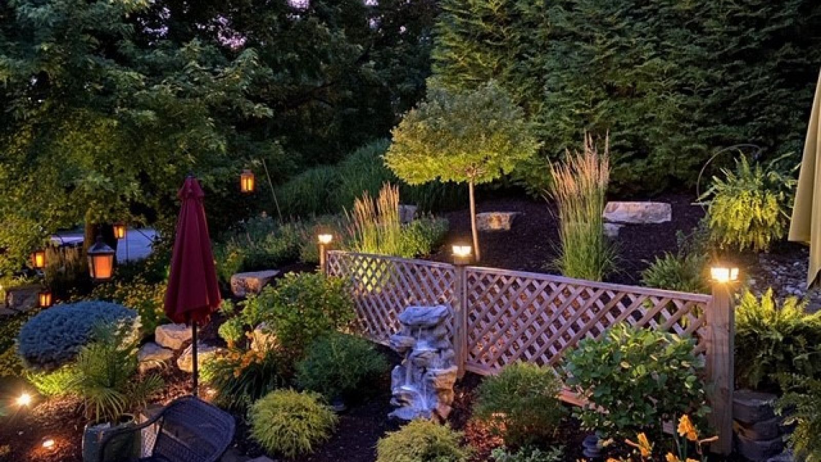 A landscaped garden at twilight featuring a lattice fence, lush greenery, small decorative lights, and a red umbrella.