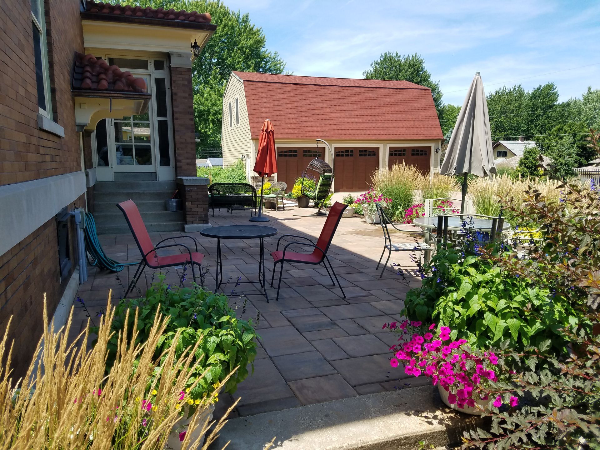 A patio featuring red chairs and a table, with a garden, umbrella, and a large shed in the background on a sunny day.