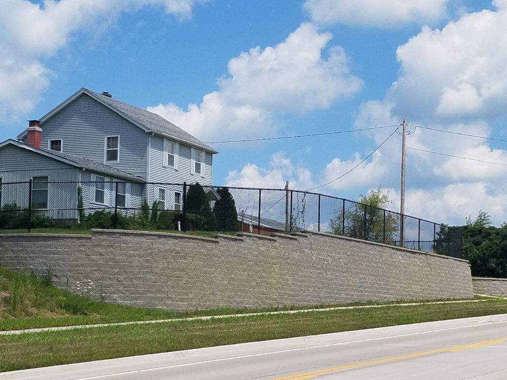 A light gray two-story house sits atop a tall, curved retaining wall made of textured stone blocks, bordered by a road.