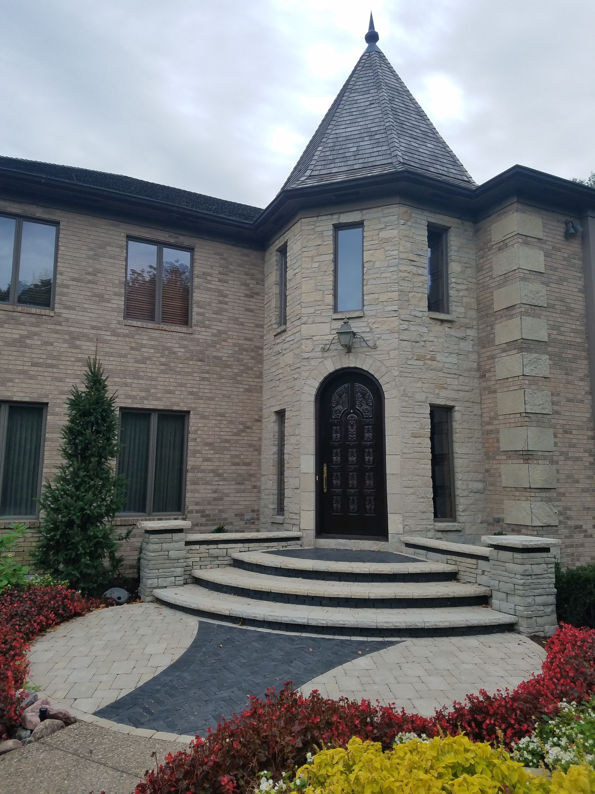 A multi-story beige stone home with a turret, arched dark wooden door, curved stone steps, and landscaped front garden.