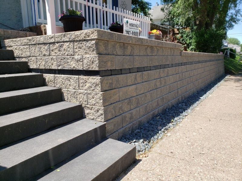 Concrete stairs lead up to a beige stone retaining wall with a decorative dark accent strip, next to a paved sidewalk.