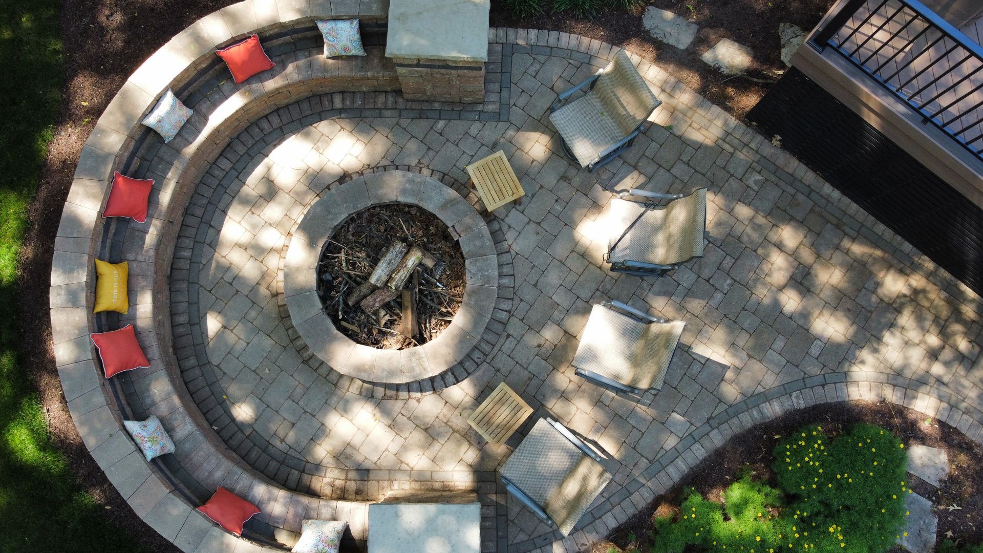 An aerial view of a stone patio with a circular fire pit, built-in curved seating with colorful cushions, and armchairs.