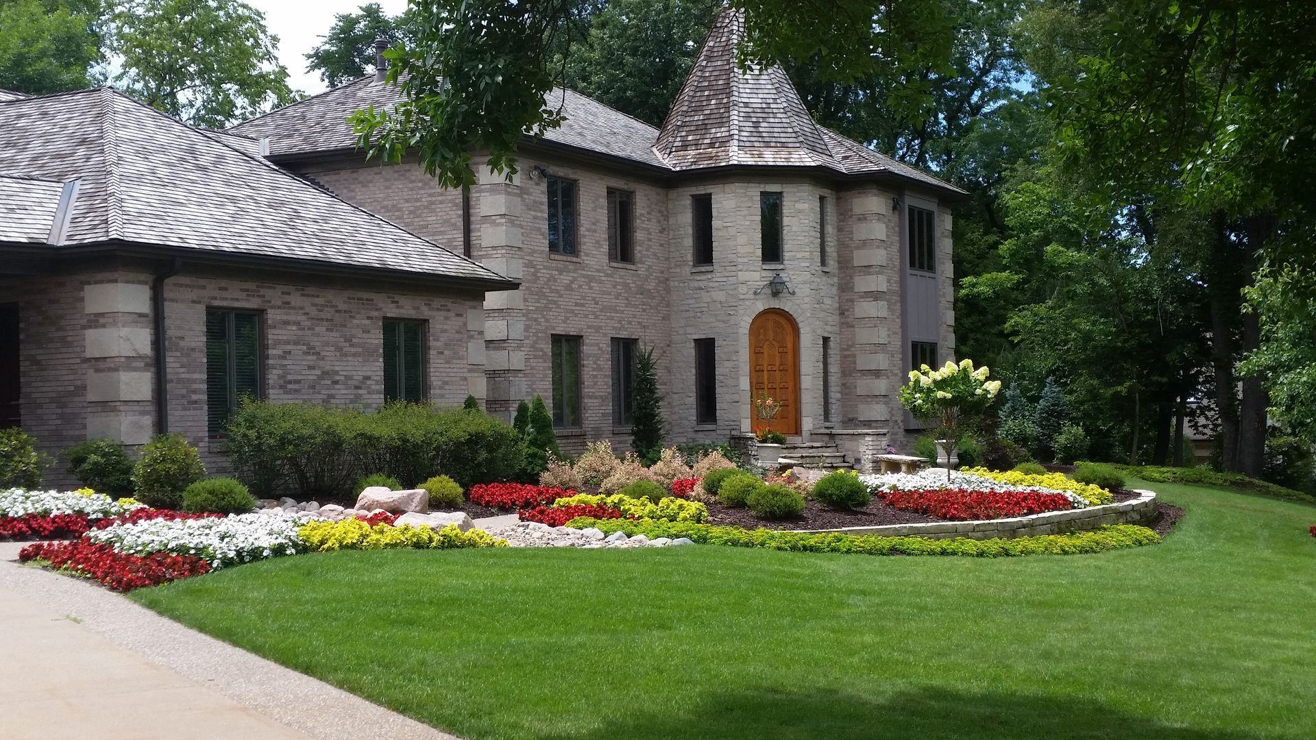 A large, two-story stone house with a turret entrance and a lush front garden featuring colorful flowers and green lawn.