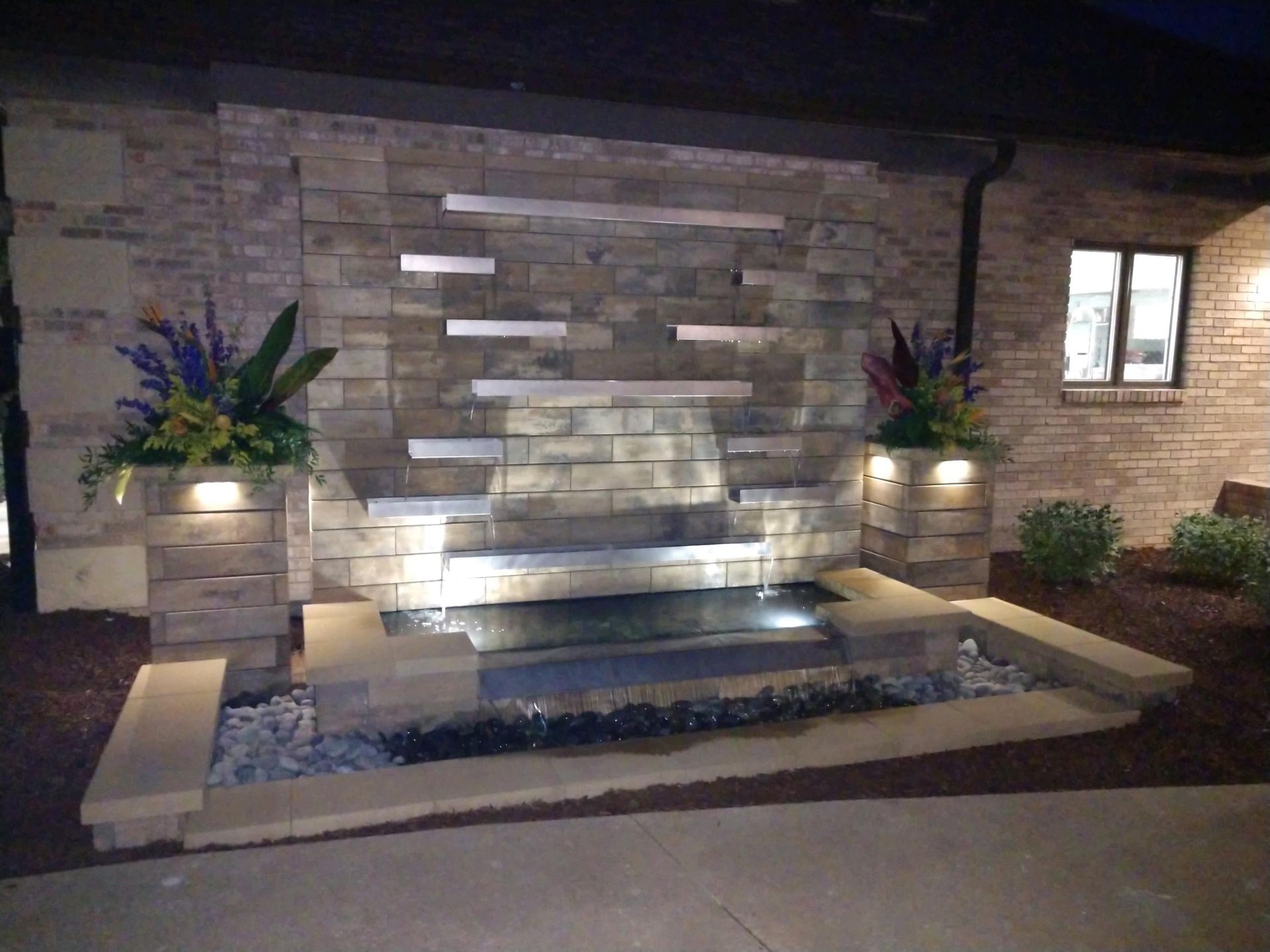 A stone wall fountain with metal tiers and water cascading into a dark basin, framed by planters and outdoor lighting.