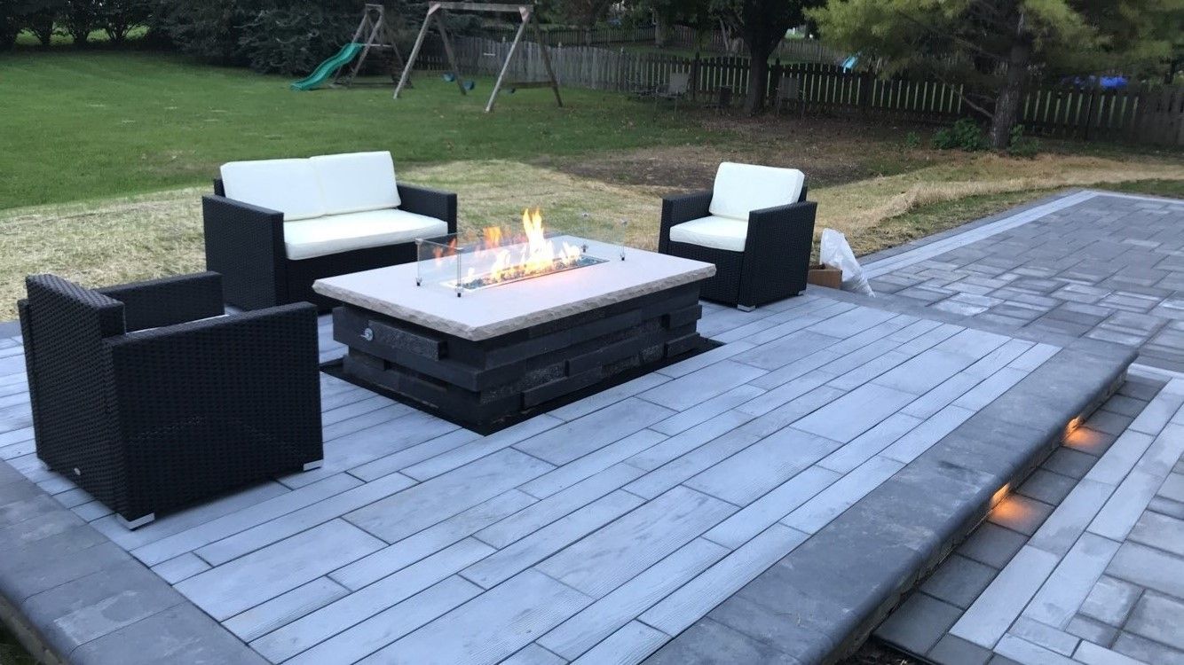 A stone patio with a lit fire pit, a loveseat, two chairs with white cushions, and built-in landscape lighting at night.