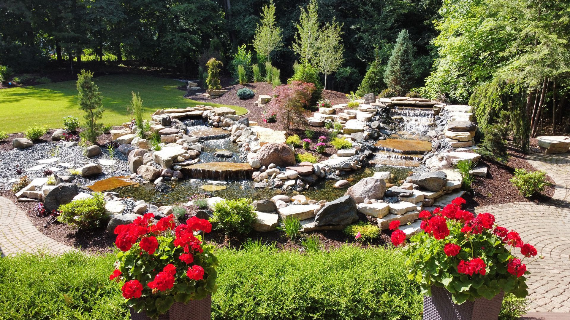 A tiered rock waterfall and pond garden with lush greenery, stone paths, and two pots of red flowers in the foreground.