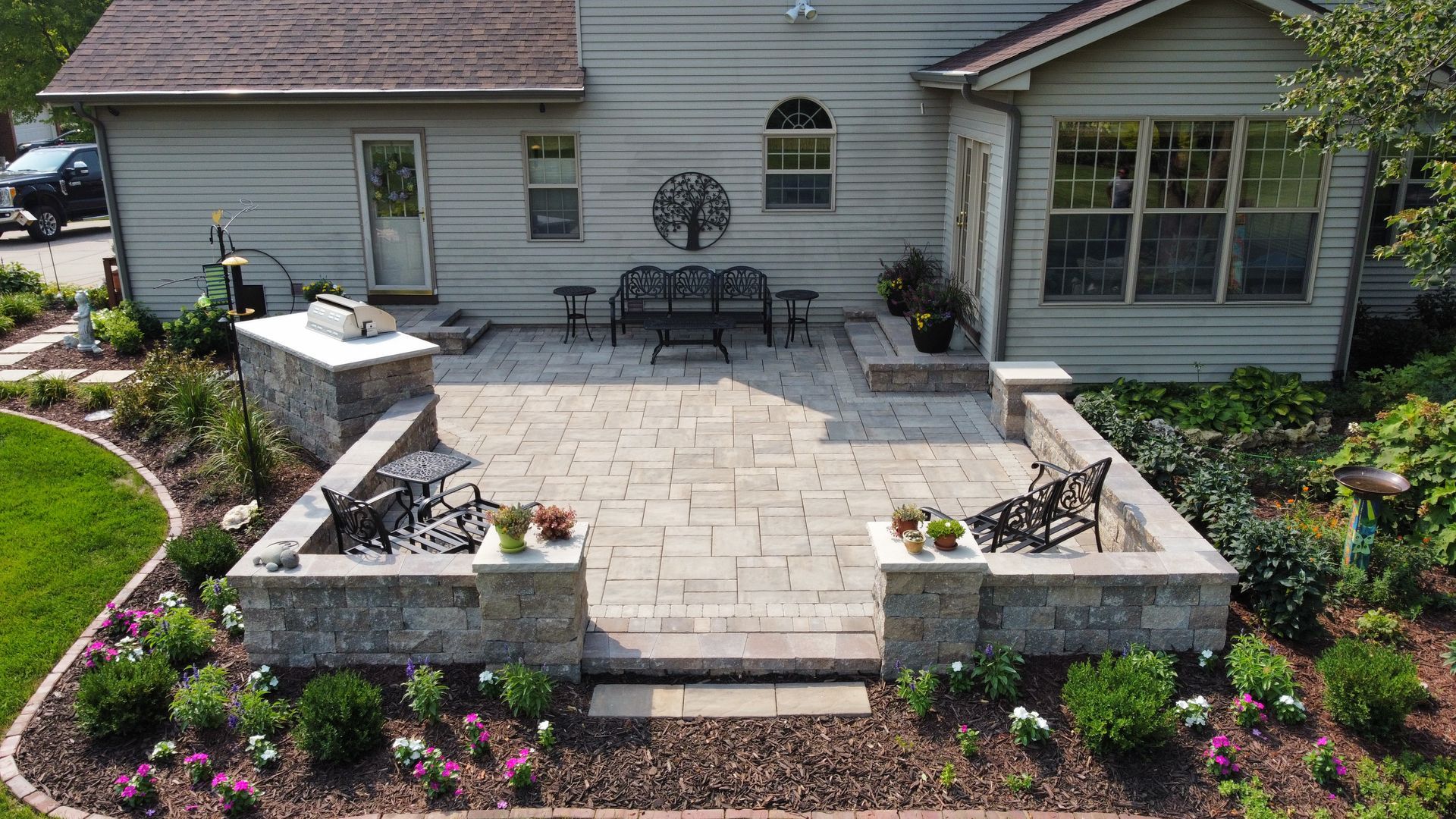 A backyard patio featuring stone pavers, a low retaining wall with pillars, outdoor seating, and surrounding landscaping.