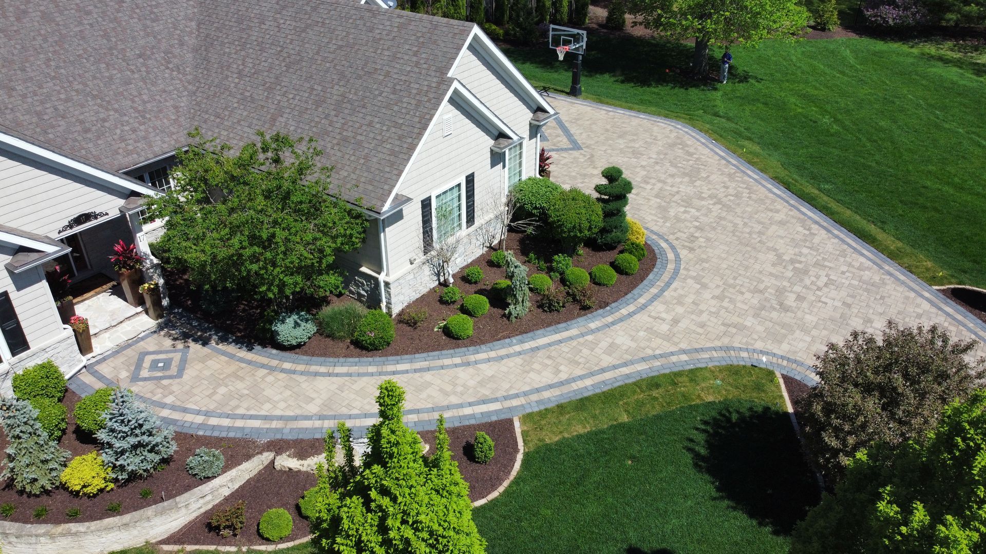An aerial view of a house featuring a curved paved driveway and walkway, bordered by landscaped gardens and a green lawn.
