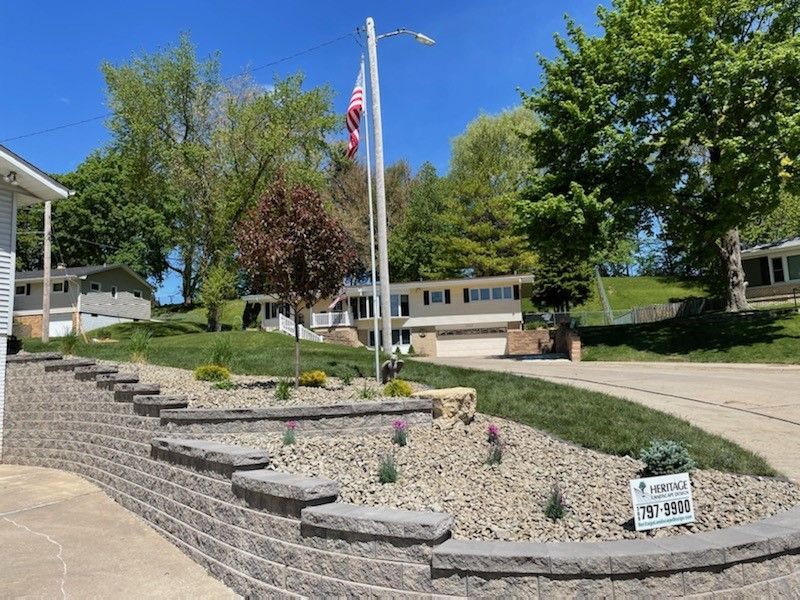 A terraced retaining wall made of gray blocks curves in front of a landscaped lawn, a house, and a flagpole under a blue sky.