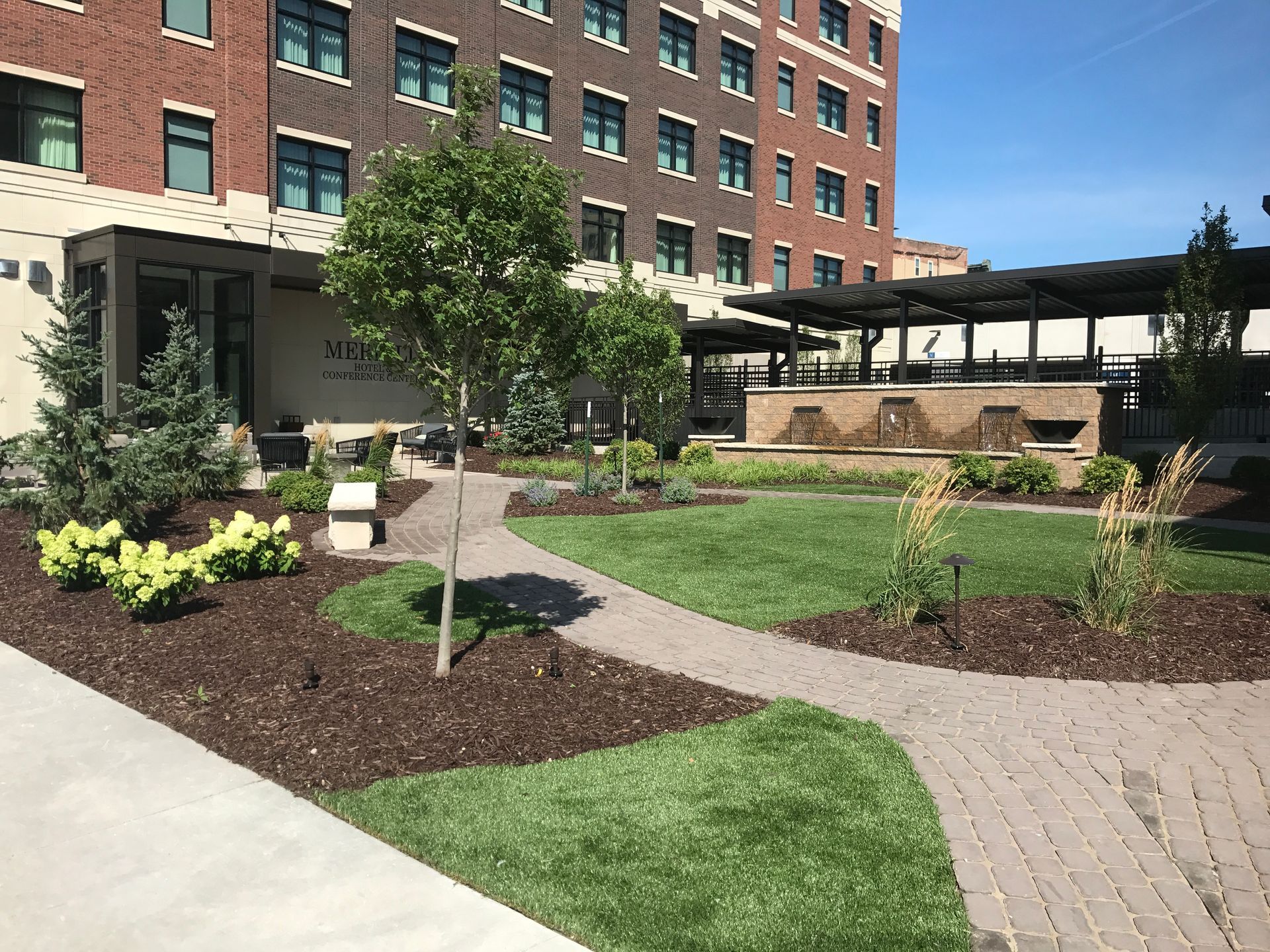 A paved walkway curves through a hotel courtyard with manicured green lawns, young trees, and a shaded outdoor lounge area.