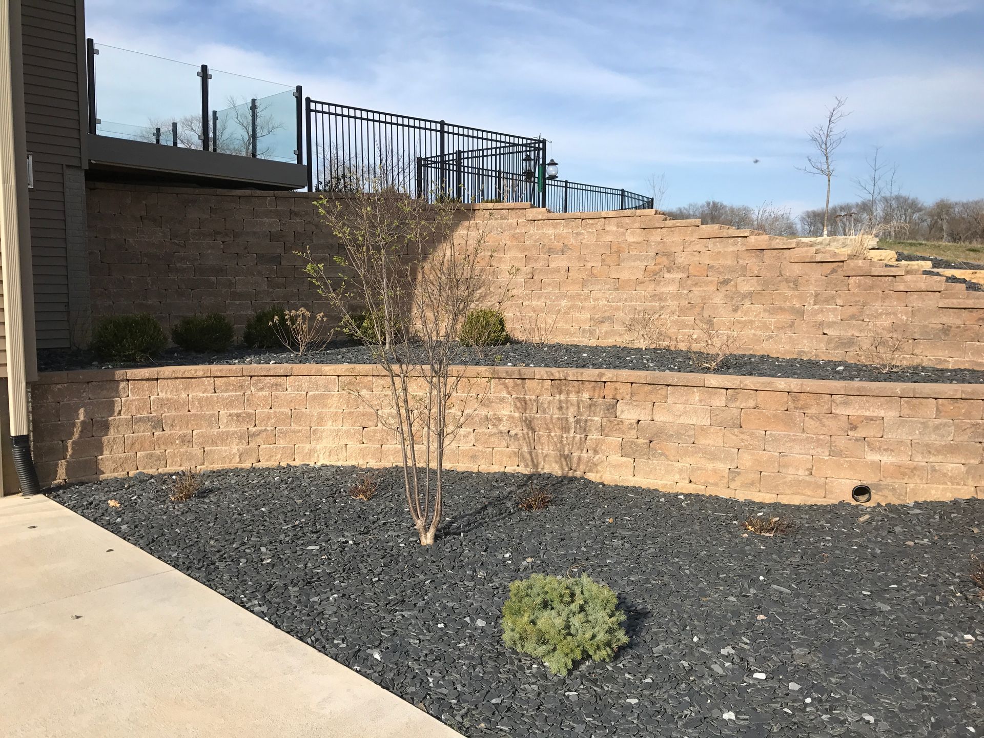 Multi-tiered tan stone retaining walls with dark gravel landscaping and a few small shrubs under a blue sky.