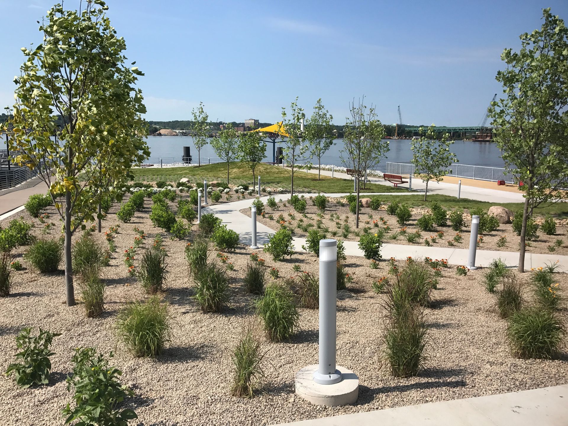 A landscaped garden with small shrubs, trees, and path lights next to a waterfront with a small beach under a clear sky.