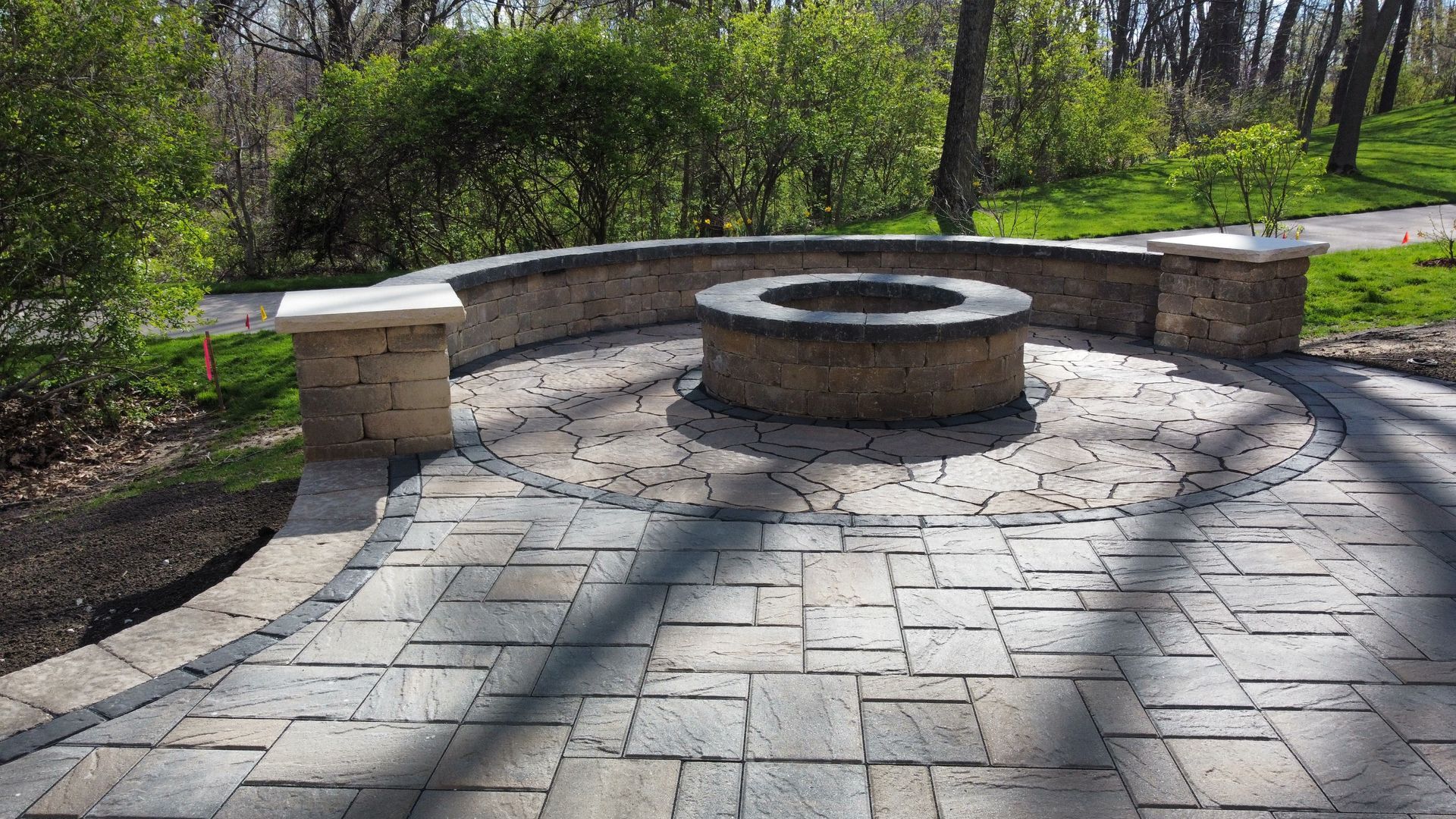A circular stone fire pit sits on a patio made of rectangular gray pavers, with a matching curved retaining wall behind it.