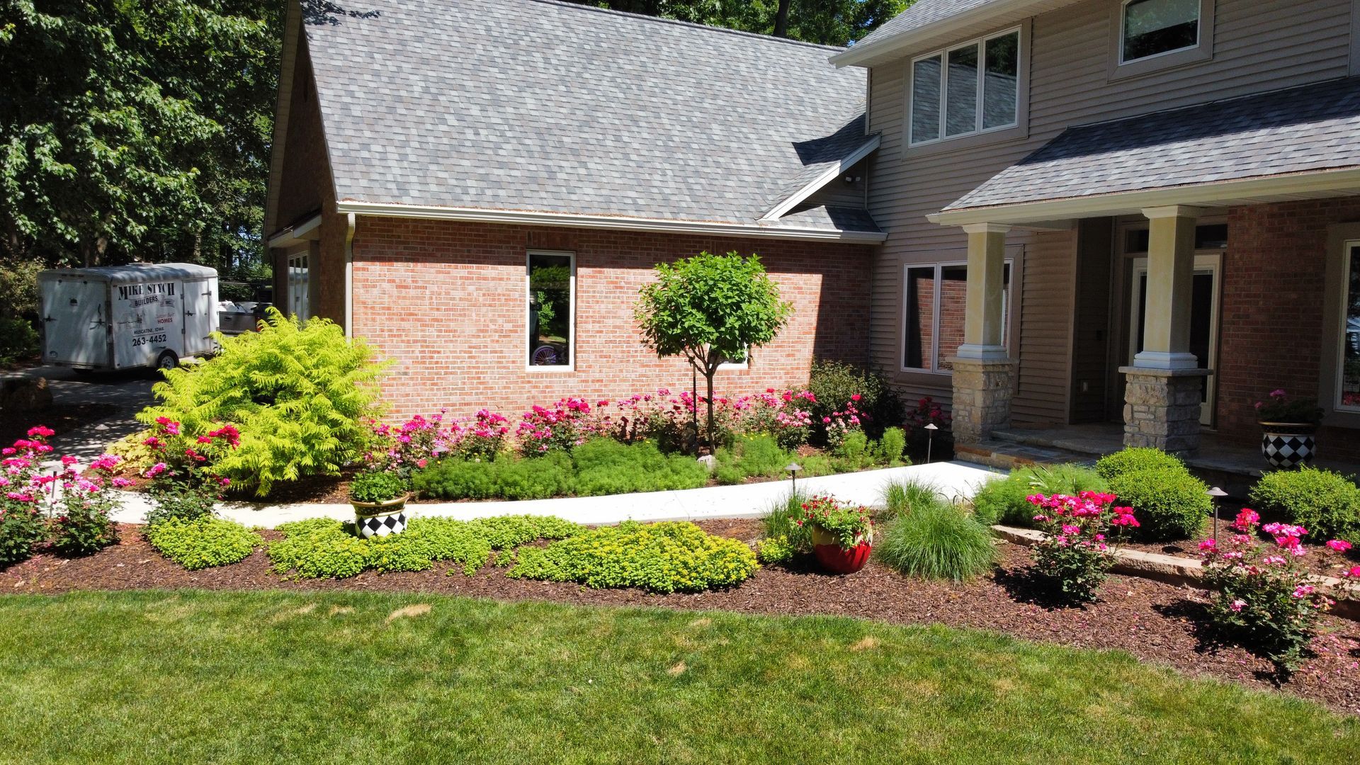 A brick house with a front porch, a pathway, and lush landscaping including shrubs, a small tree, and pink flowers.