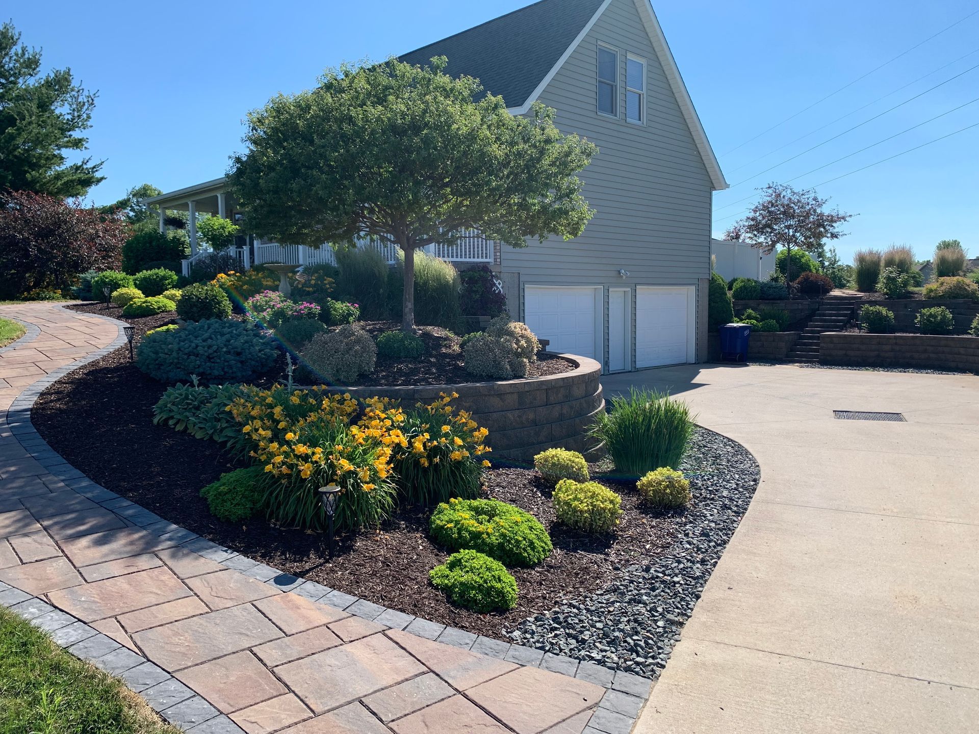 A tan house with a garage, fronted by a paved walkway and a landscaped retaining wall with trees, shrubs, and flowers.
