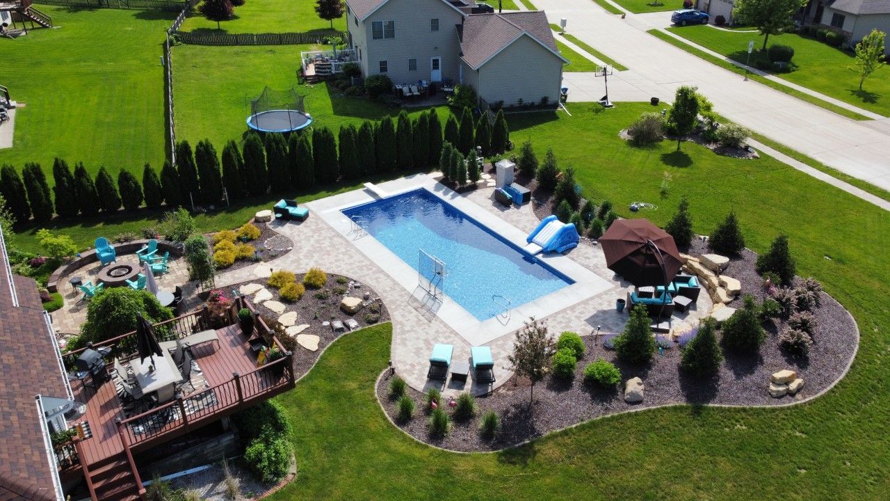 Aerial view of a rectangular backyard pool, patio area, fire pit, and wooden deck on a sunny day.