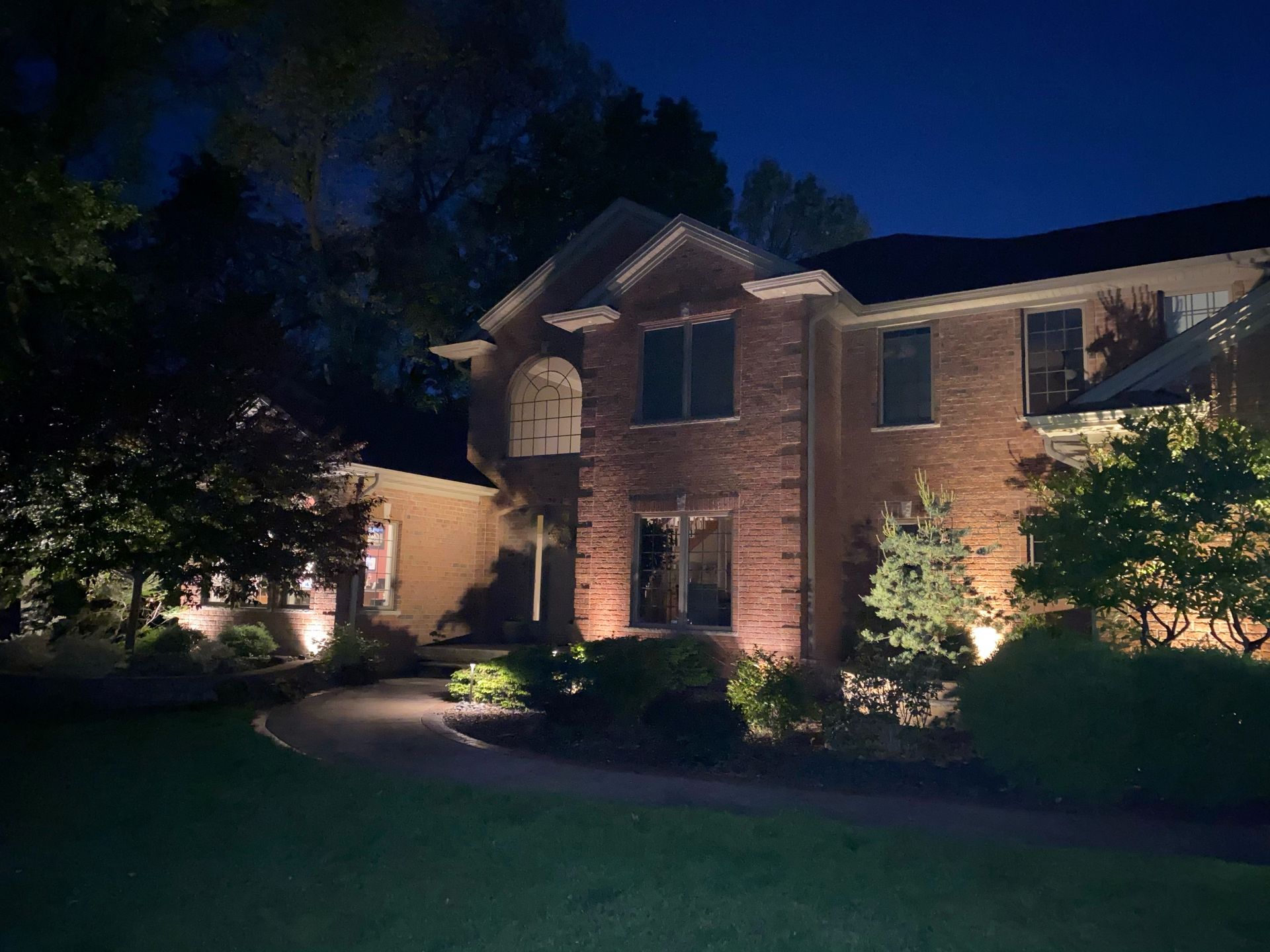 A two-story brick house at night, illuminated by warm outdoor landscape lighting along the walkway and front garden.