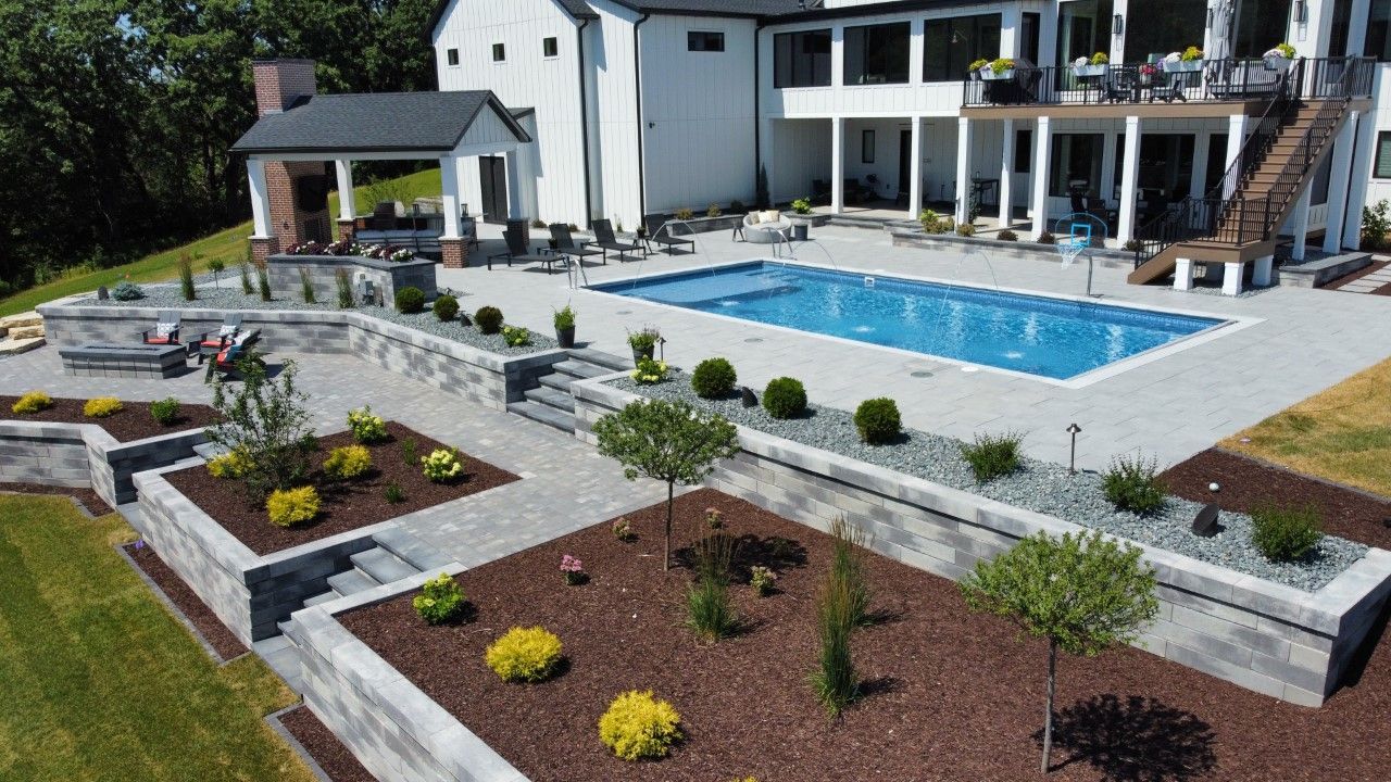A modern house with a pool, a covered patio with an outdoor fireplace, and tiered landscaping with shrubs and mulch.