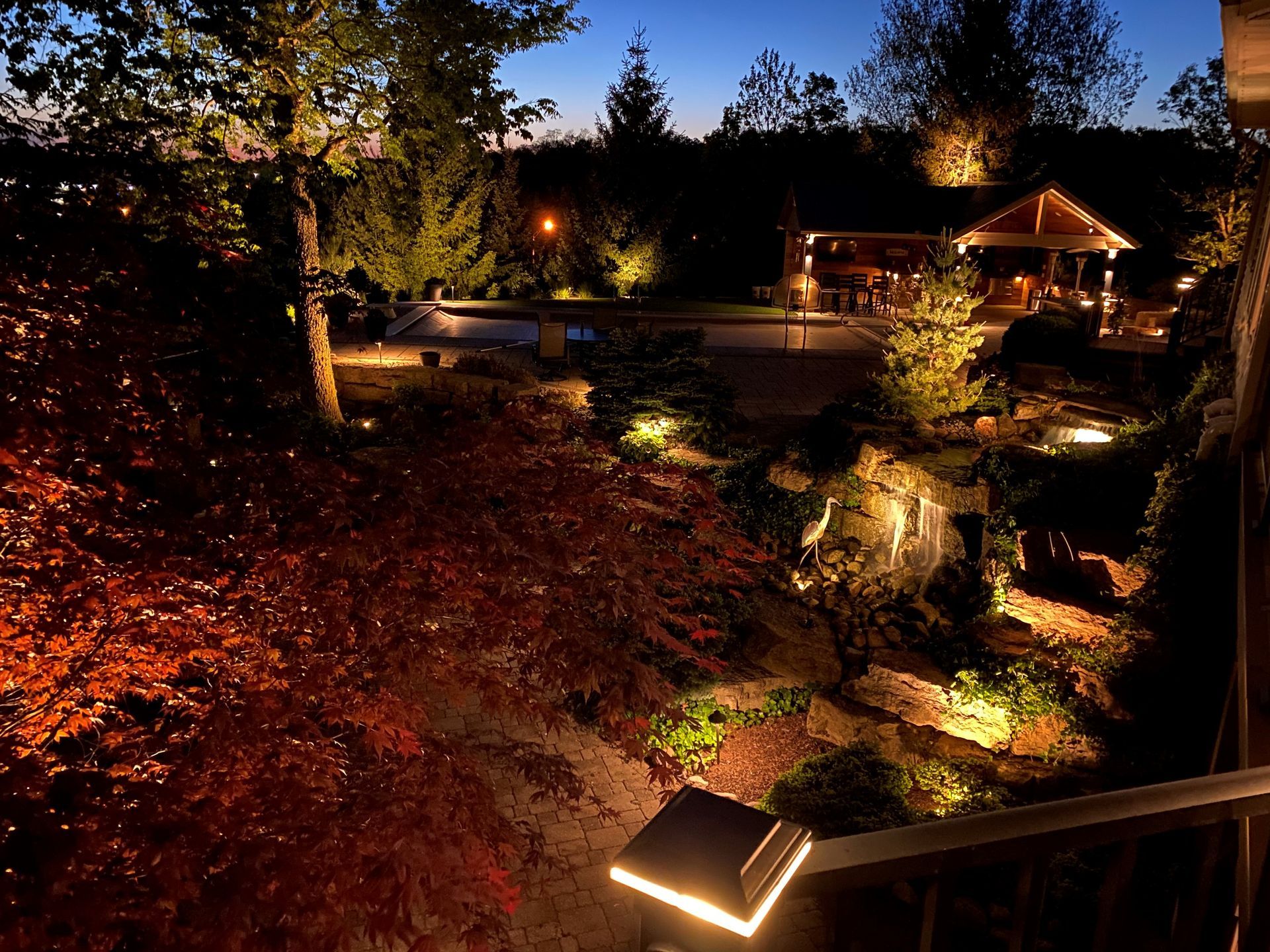 An illuminated backyard garden at dusk with a pool and a gazebo in the background, viewed from an elevated deck.