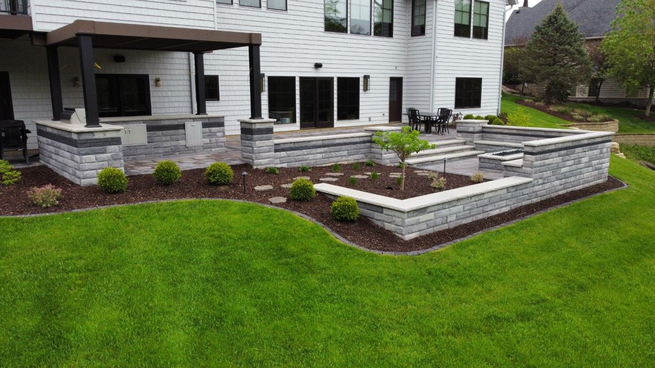 A multi-level stone patio with a pergola and outdoor kitchen in a landscaped backyard with a lush green lawn.