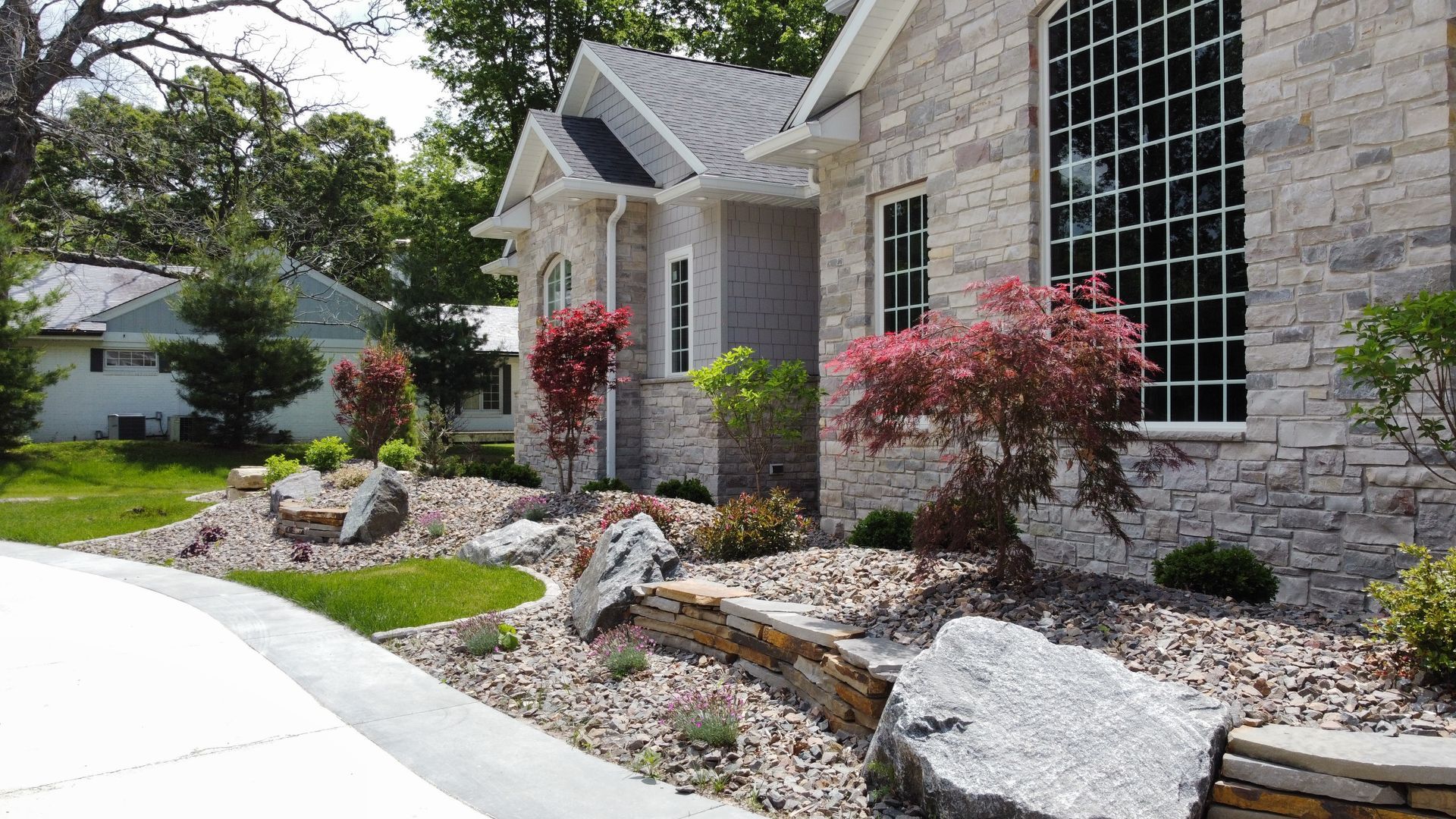 A stone house with landscaped front yard, including a low retaining wall, large rocks, and small trees and shrubs.