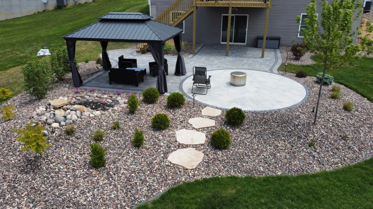 A backyard landscape featuring a dark gazebo with patio furniture, a circular stone fire pit area, and a stone path.