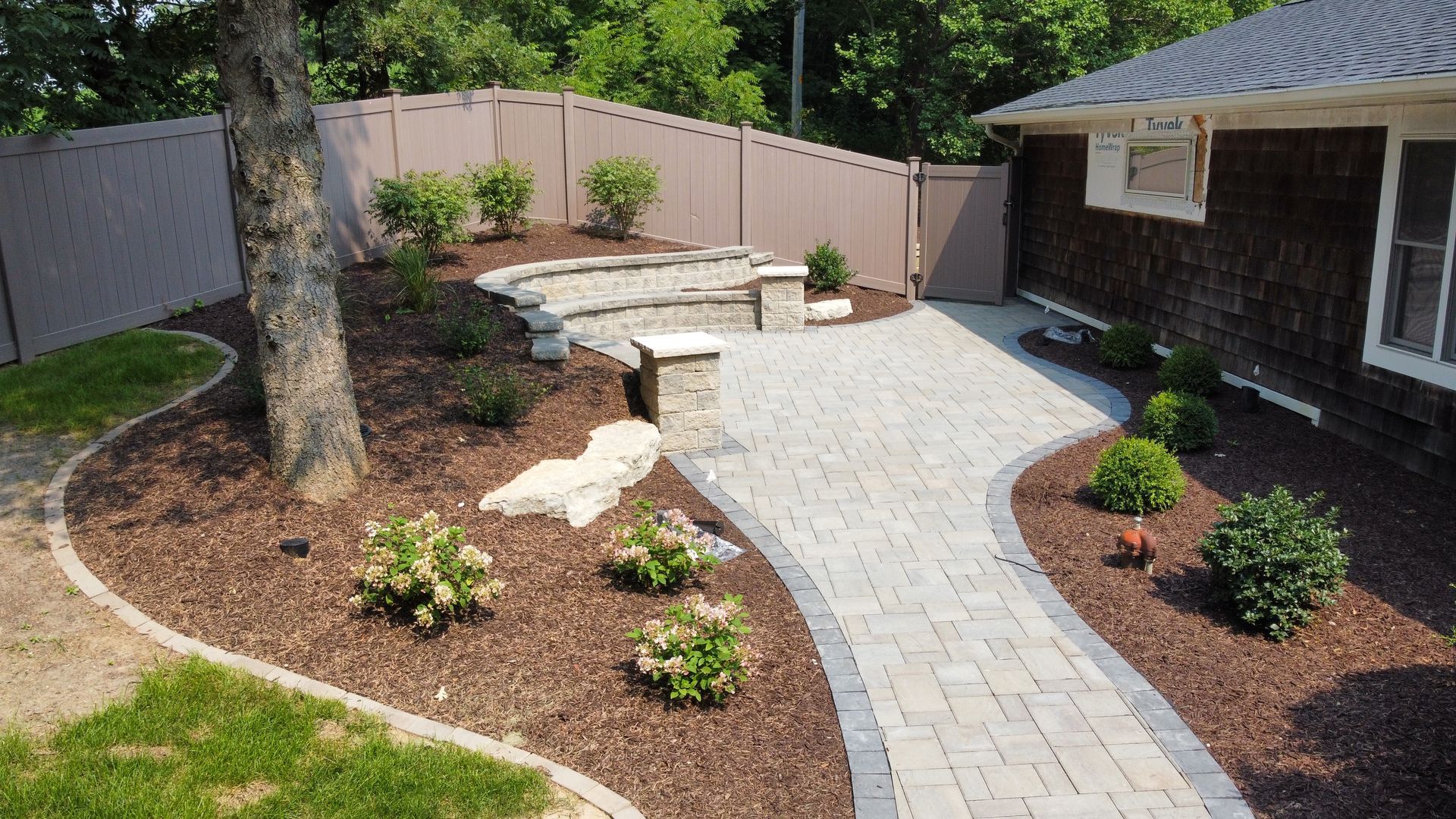 A stone paver walkway leads to a curved stone bench in a landscaped backyard with mulch beds, shrubs, and a tree.