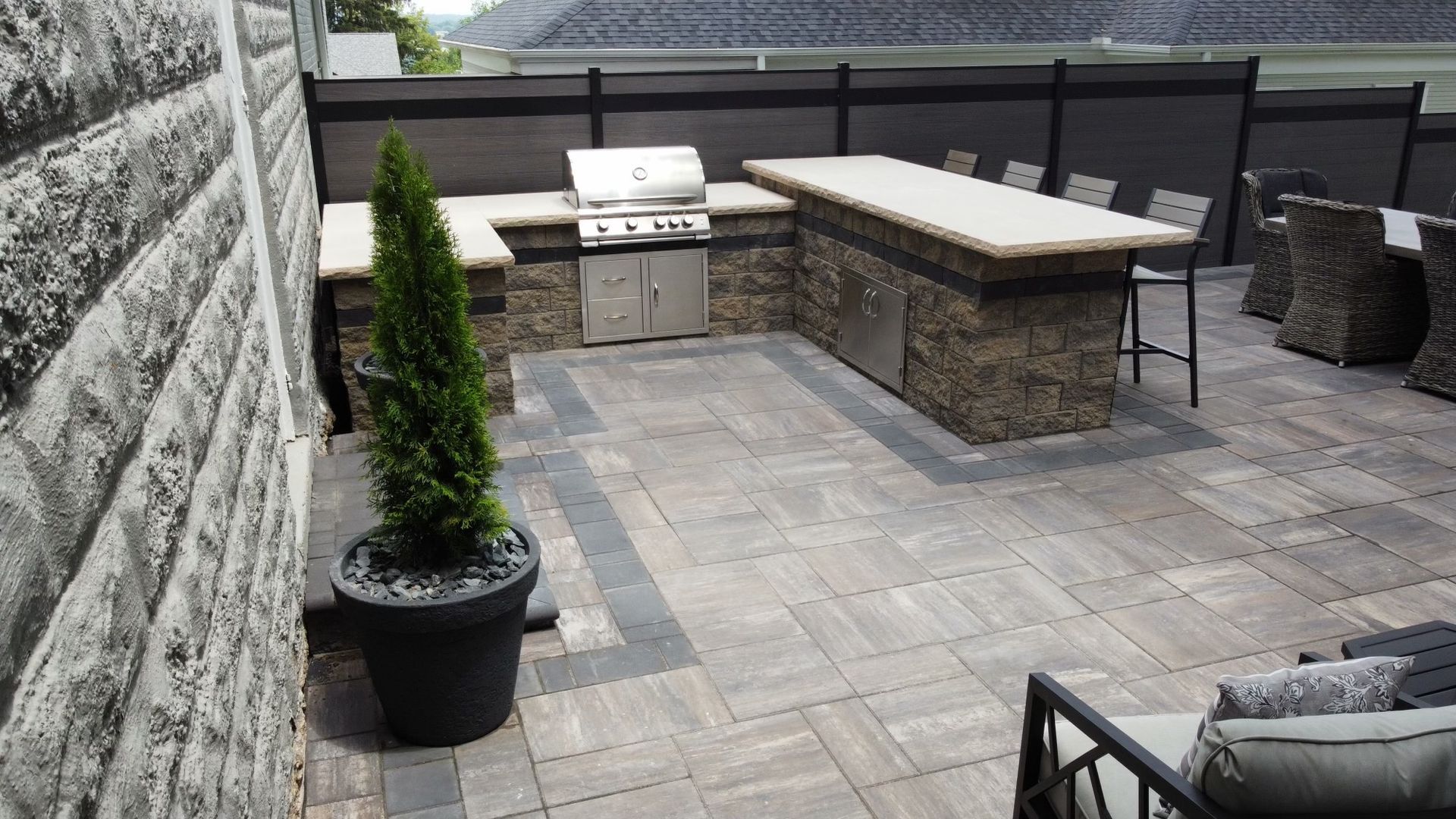 Outdoor patio featuring a stone kitchen island with a built-in grill, bar stools, and a potted evergreen tree.