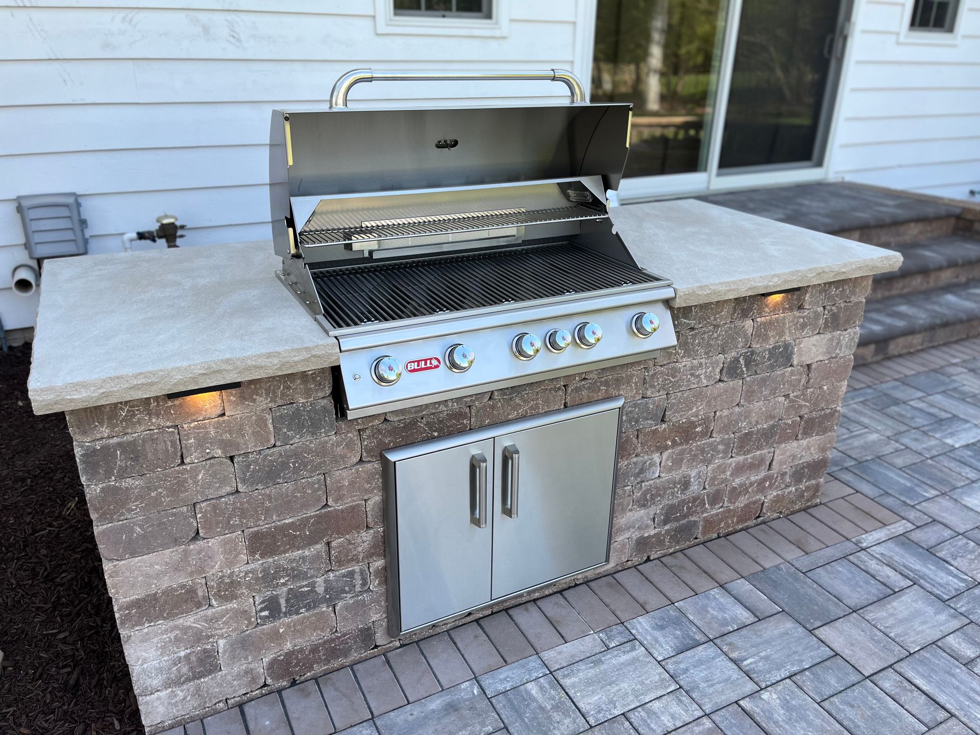 An outdoor brick grill island with a stainless steel built-in grill on a stone patio in front of a house.