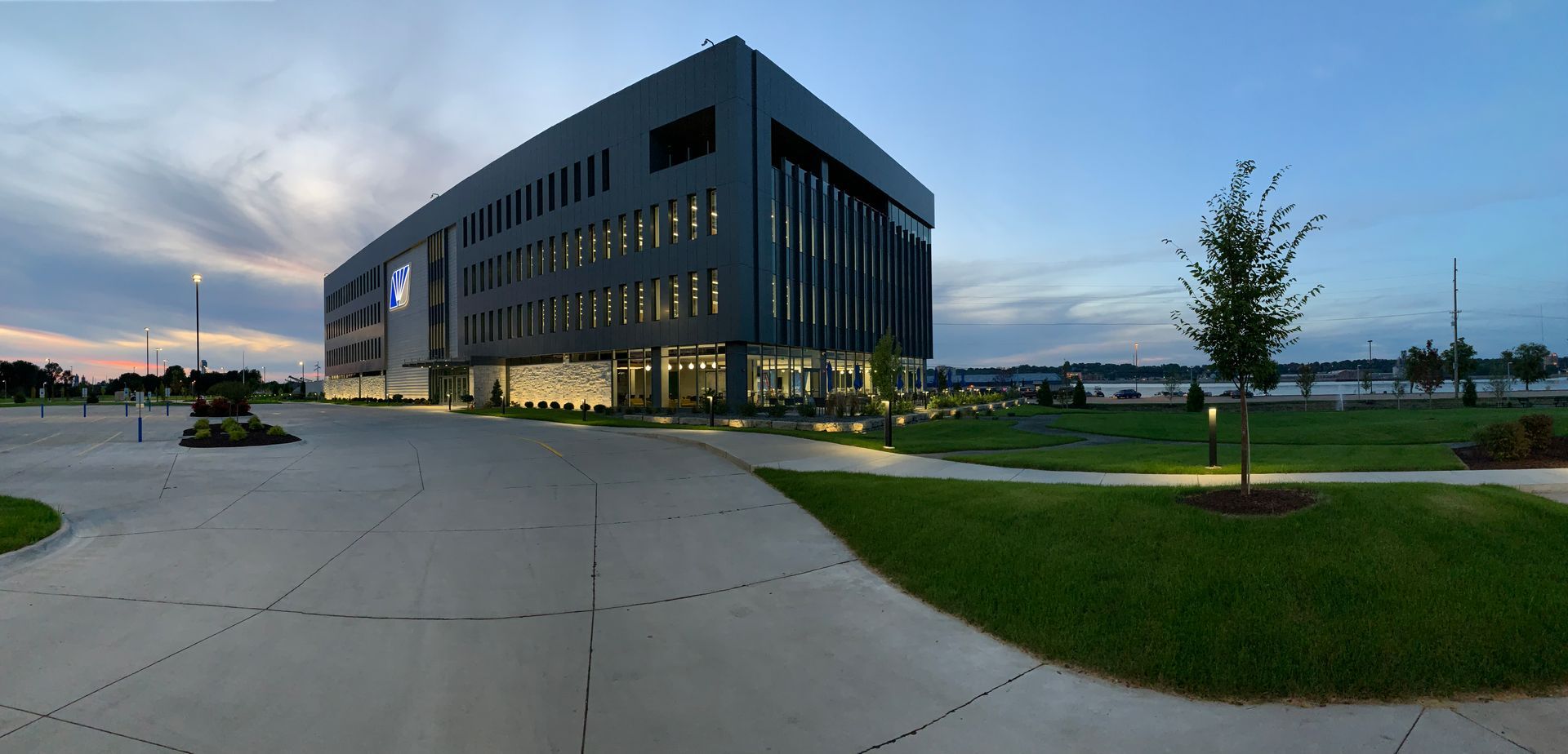 A modern, multi-story dark gray building sits beside a grassy lawn under a twilight sky.