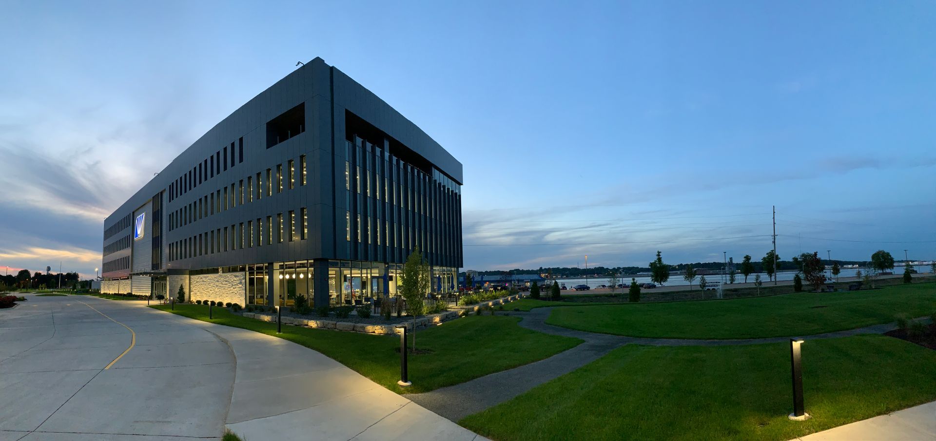 Modern, multi-story grey office building at dusk, illuminated by exterior lights, surrounded by lawns and walkways.