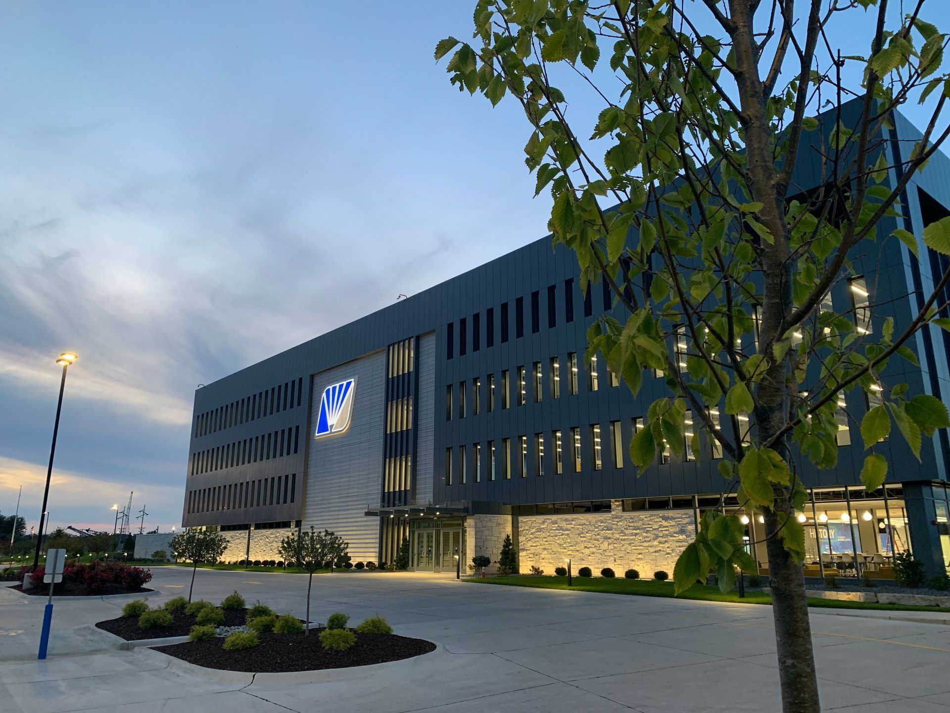 A modern, multi-story office building with a dark facade and stone-textured base at dusk, framed by a tree in the foreground.