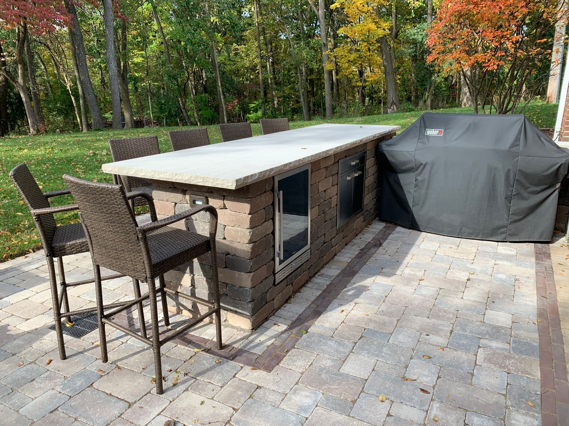 Outdoor patio with a stone kitchen island, bar stools, and a covered grill, set against a backdrop of autumn trees.
