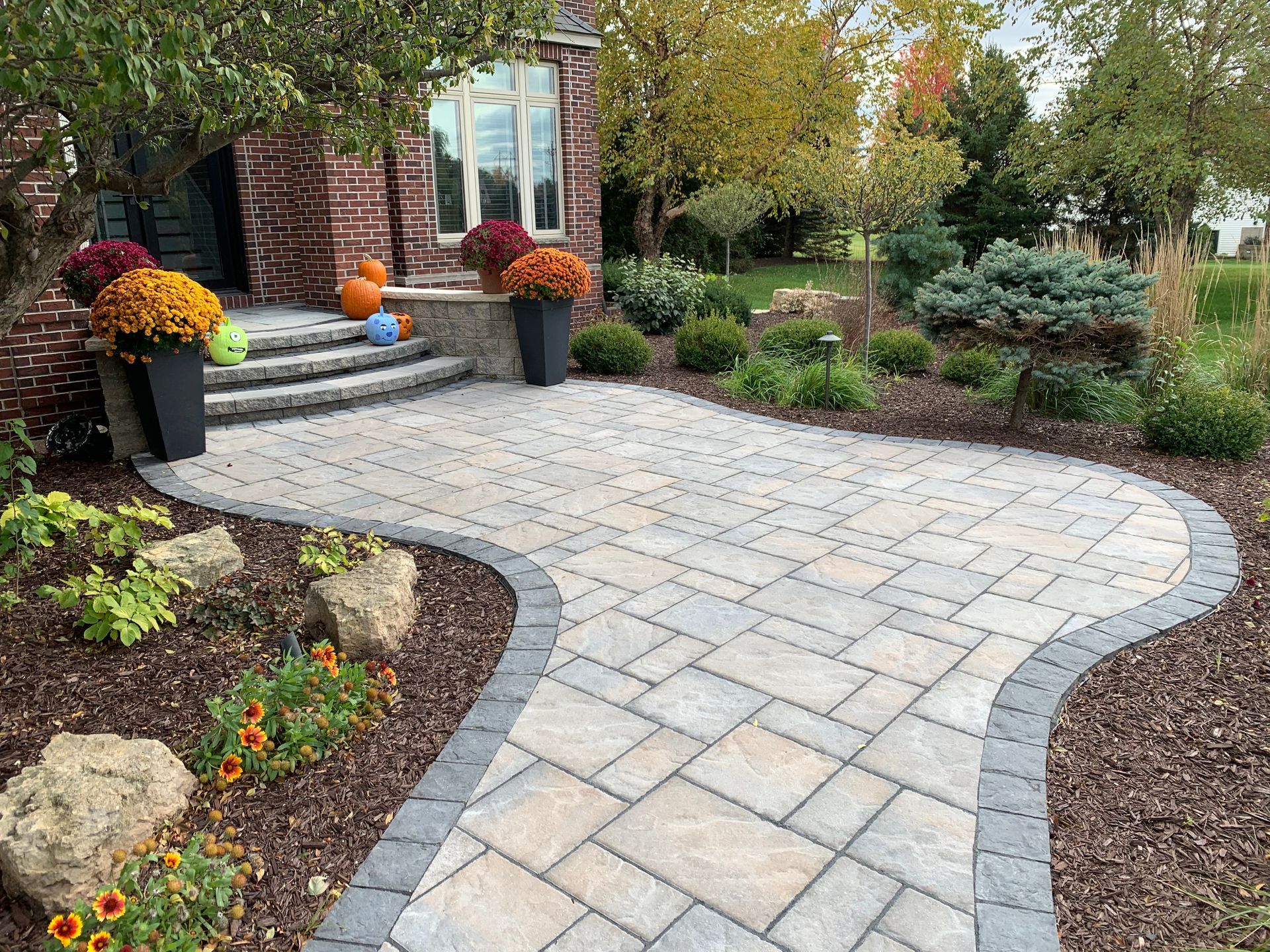 A brick house entrance with a stone paver walkway, potted mums, and landscaped gardens with mulch and decorative stones.