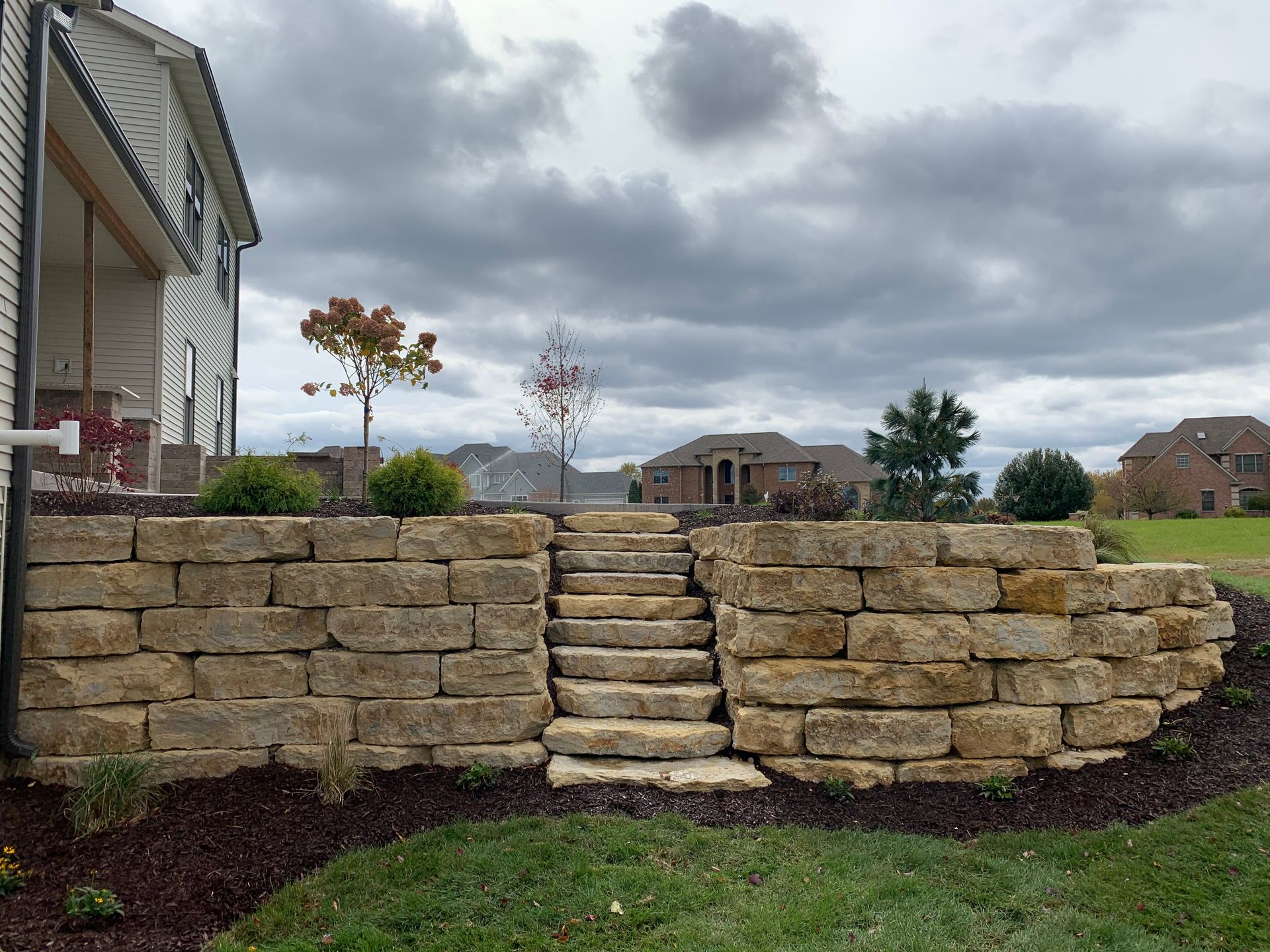 Stone retaining wall with a central set of stone steps leading up to a yard, next to a house under a cloudy sky.