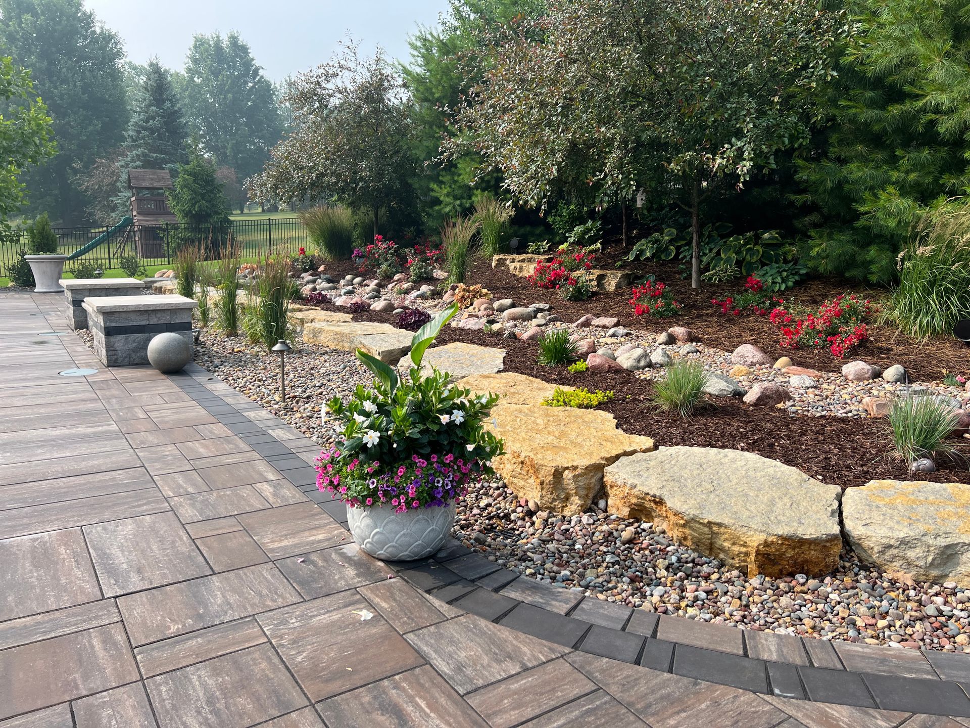 A stone patio sits next to a landscaped garden featuring large boulders, dark mulch, flowering shrubs, and a potted plant.