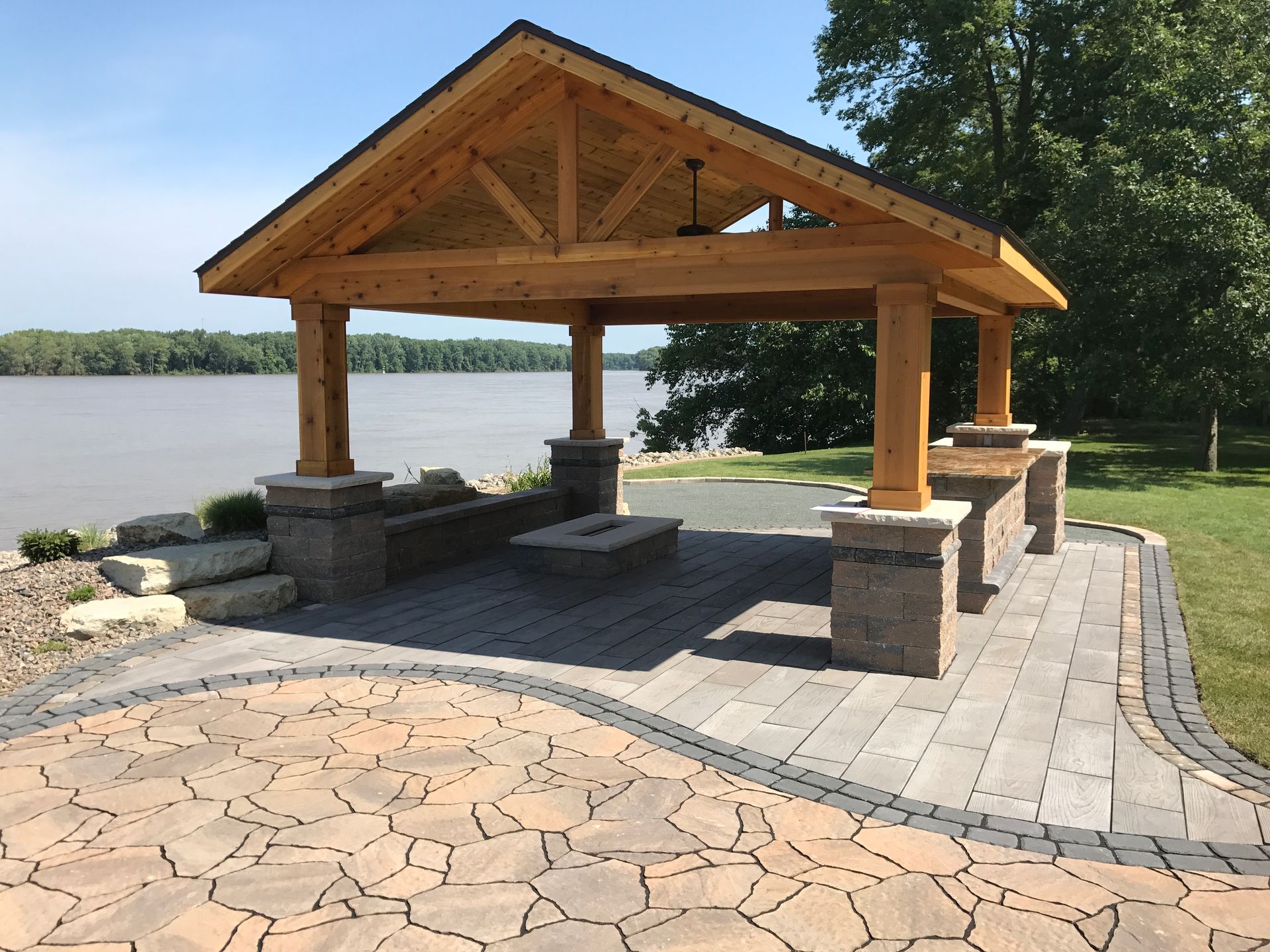 A wooden pavilion with stone pillars overlooks a river from a paved stone patio with a fire pit.