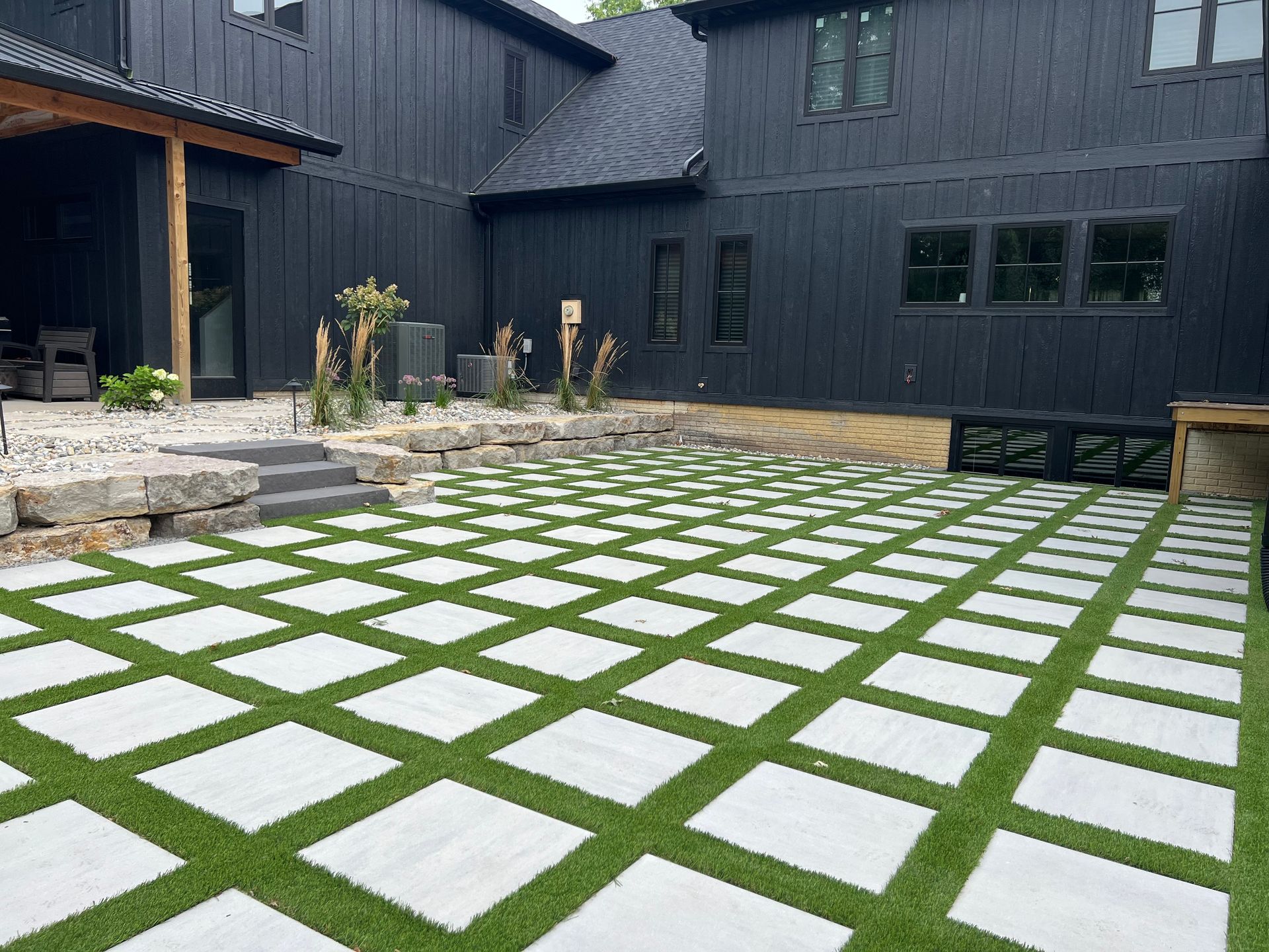 Modern black home with a grid-patterned patio of square stone pavers set in green turf leading to stone steps.