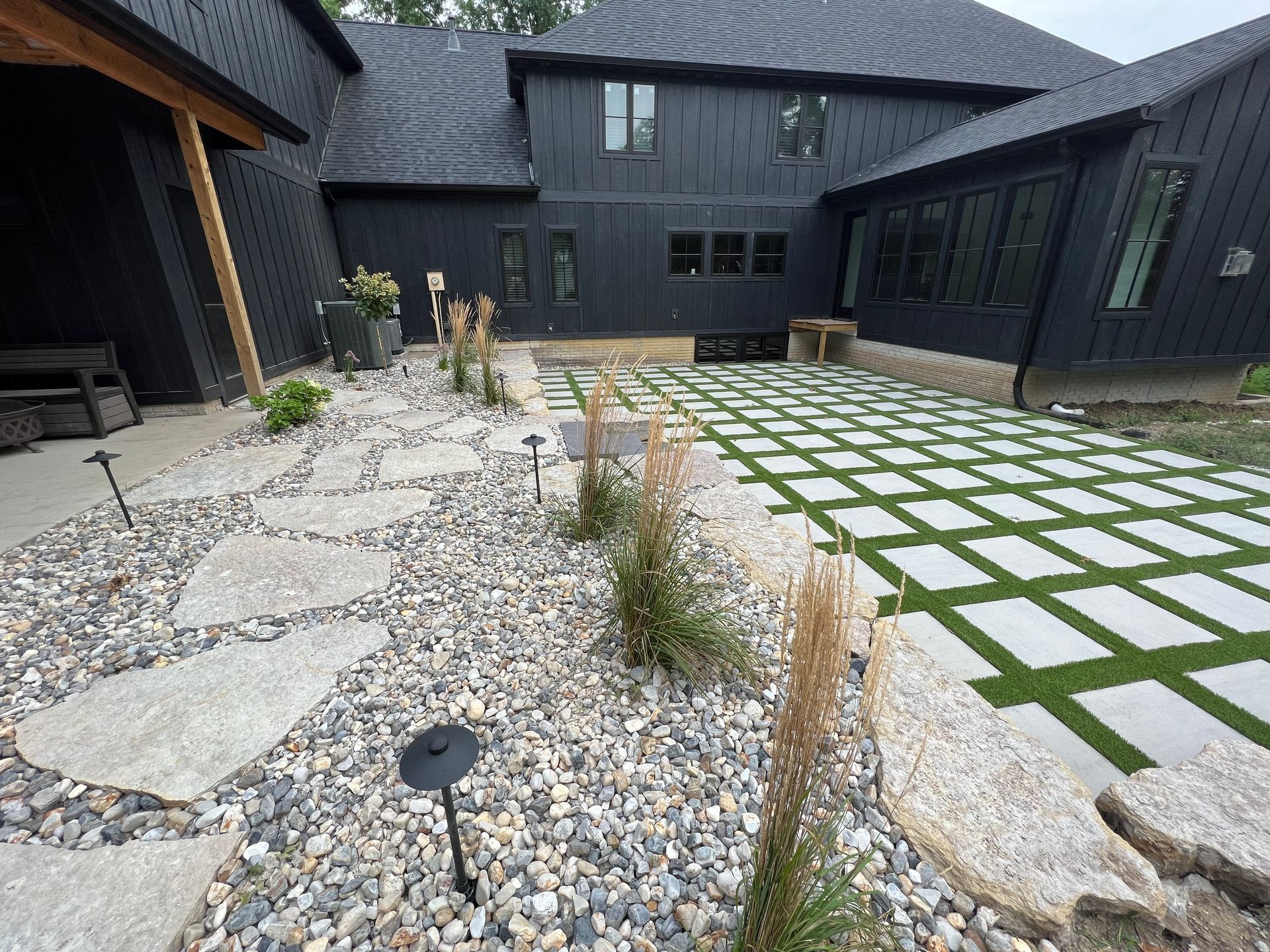 A modern black house exterior with a landscape featuring stone paths, gravel, ornamental grasses, and a grid patio.