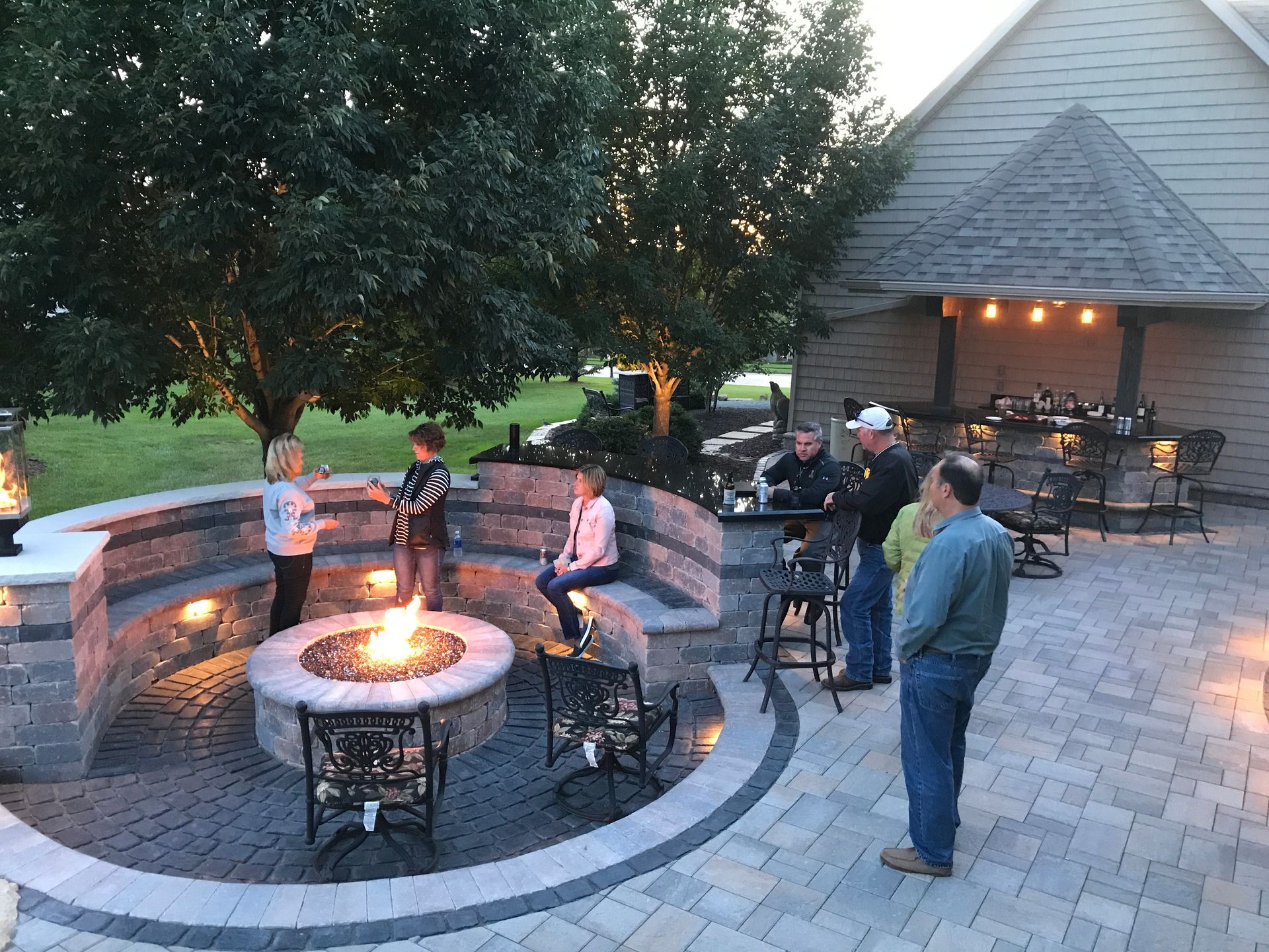 A group gathers around a glowing circular fire pit on a paved outdoor patio near a home with an outdoor bar.