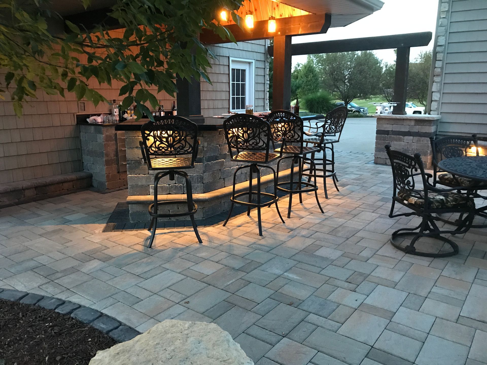 An outdoor patio features a stone bar with ornate metal stools, pendant lighting, and a fire pit area with stone pavers.